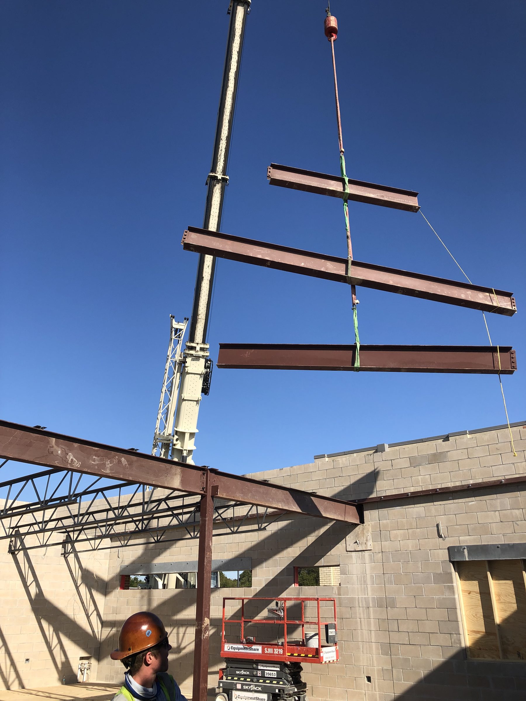 Three steel beams rigged on a single crane line, lifted above a masonry building under construction