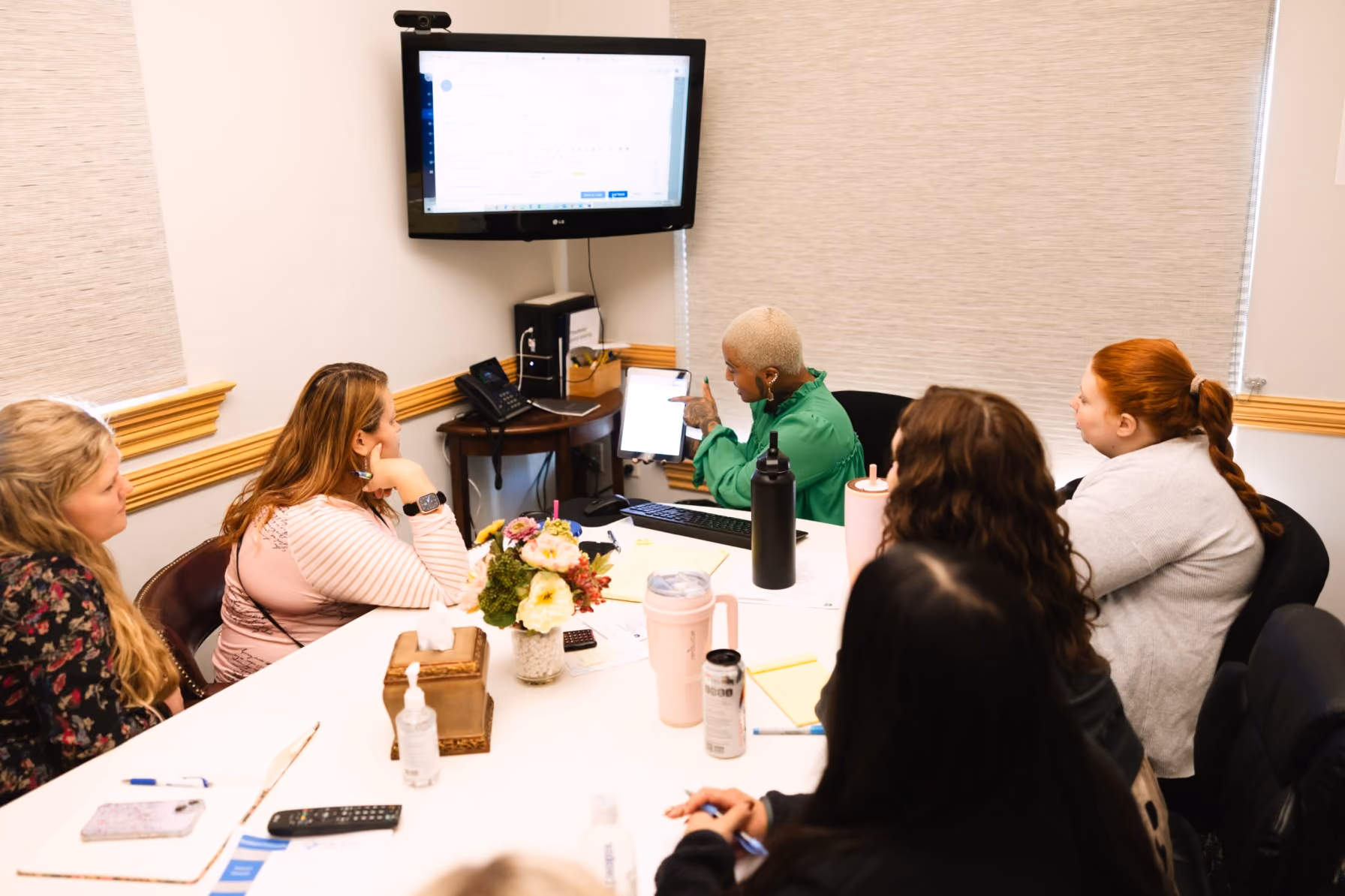 Group of people sitting at a conference table watching a presenter with a tablet and screen.