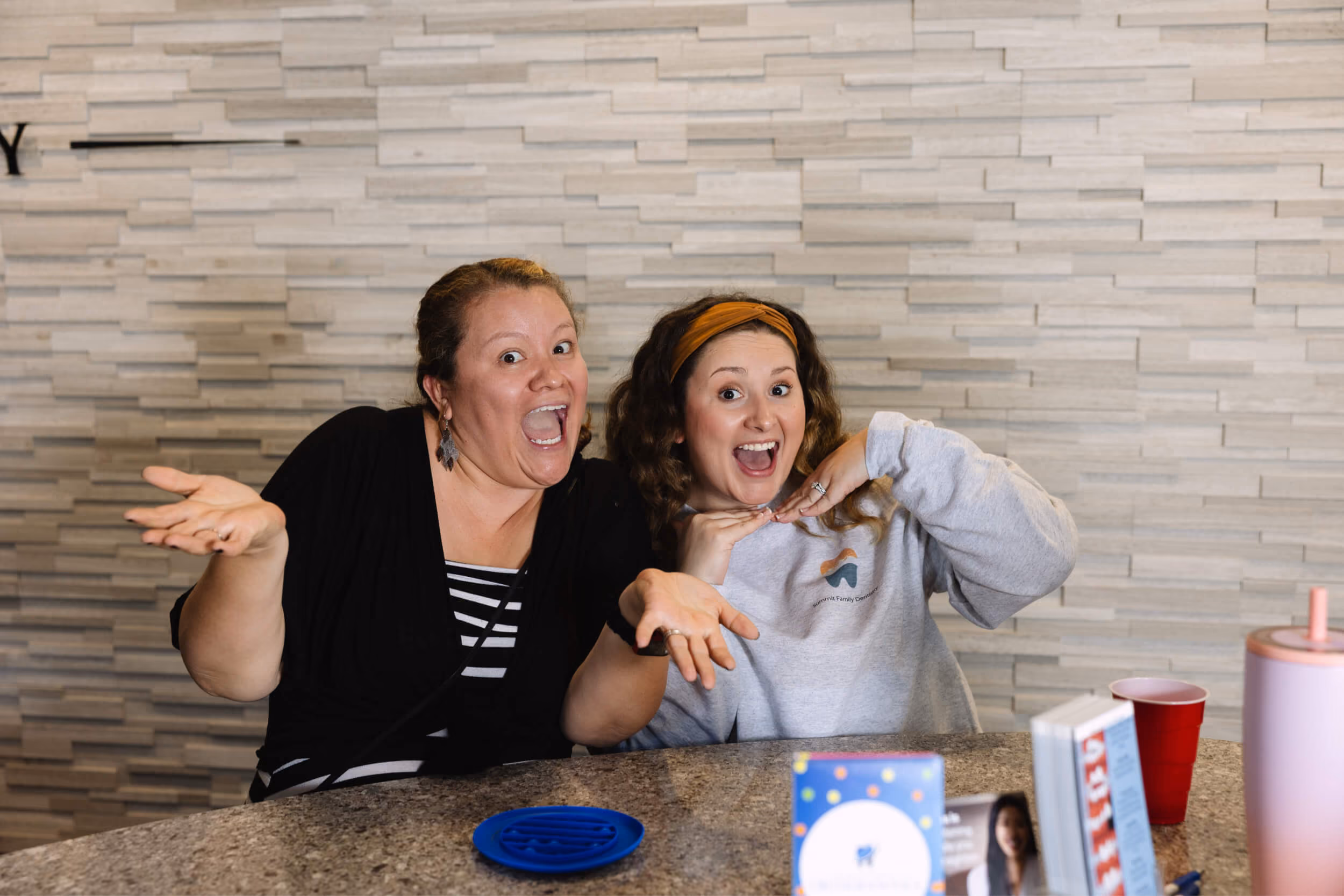 Two women sitting at a table smiling widely and posing with their hands up near their faces.