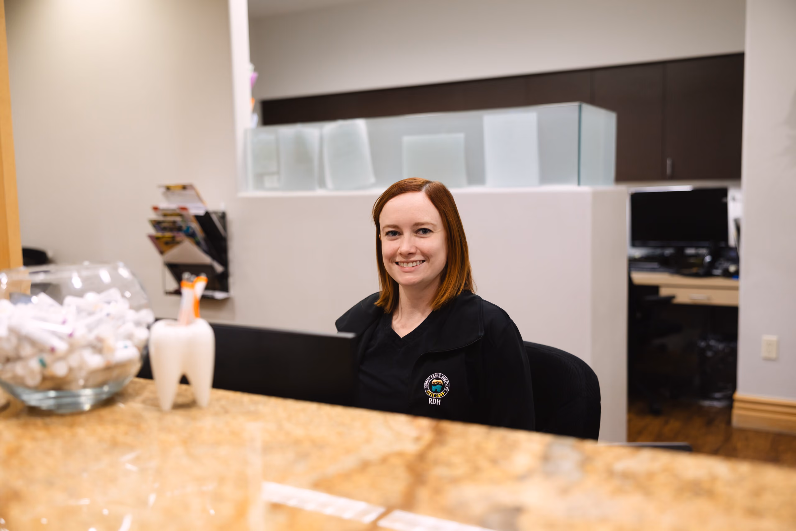 Smiling receptionist in black uniform sitting at a dental office front desk with tooth model nearby.