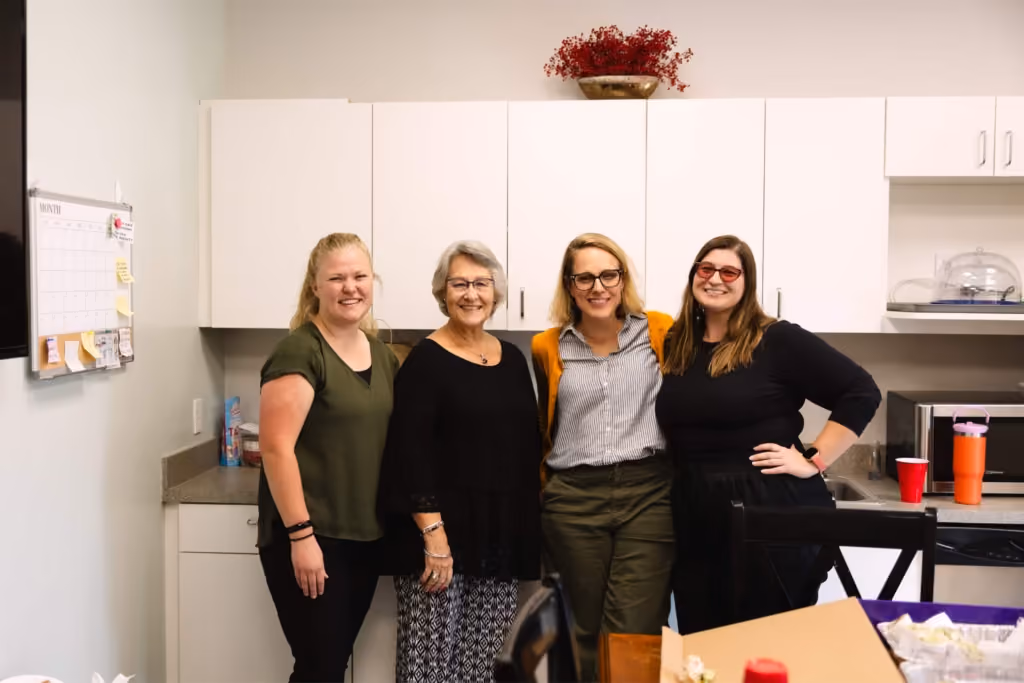 Four women smiling and standing together in a kitchen with white cabinets and a red flower pot above.