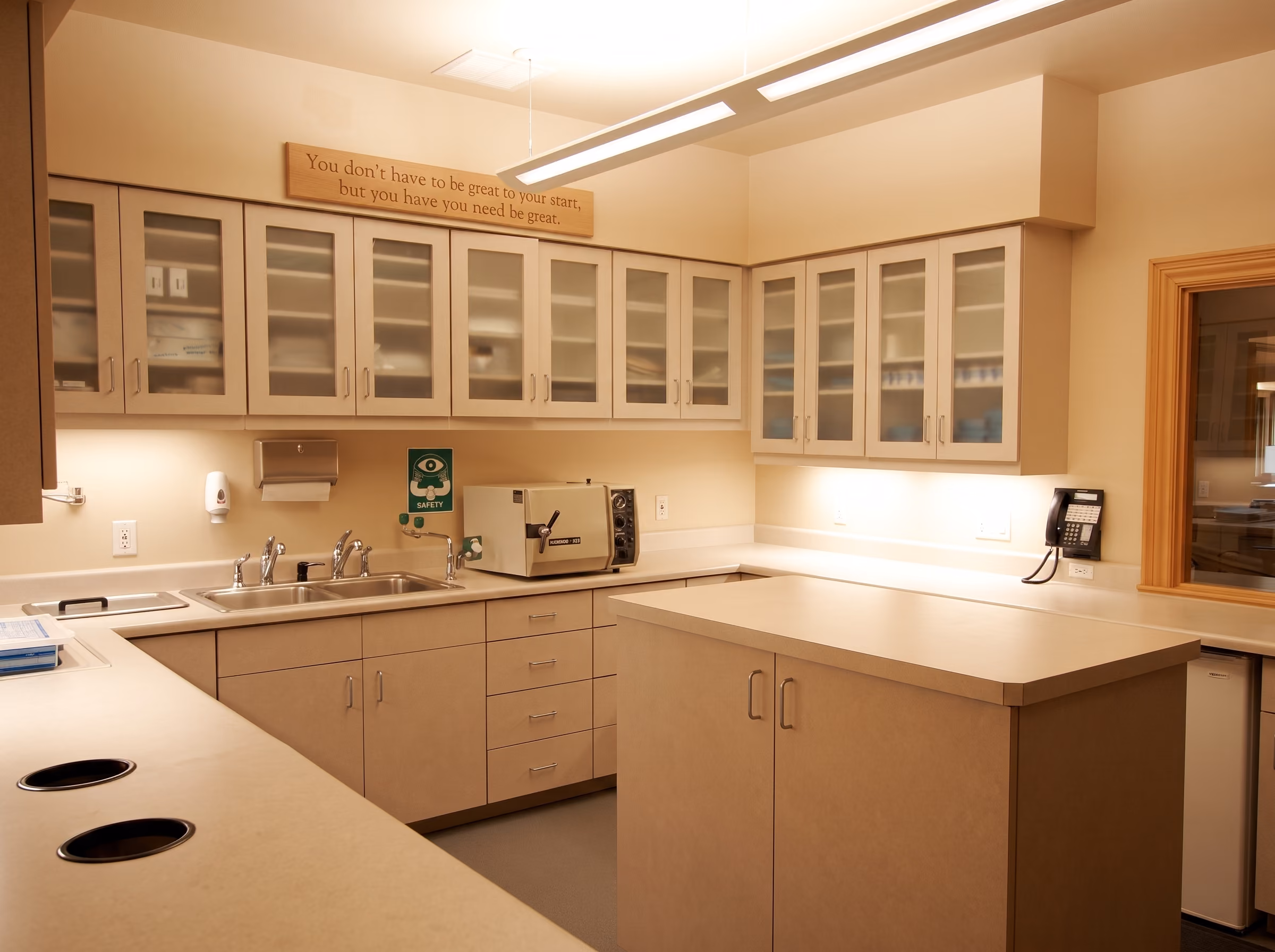 Clean beige laboratory or medical room with cabinets, sinks, sterilizer, phone, and a motivational sign above.