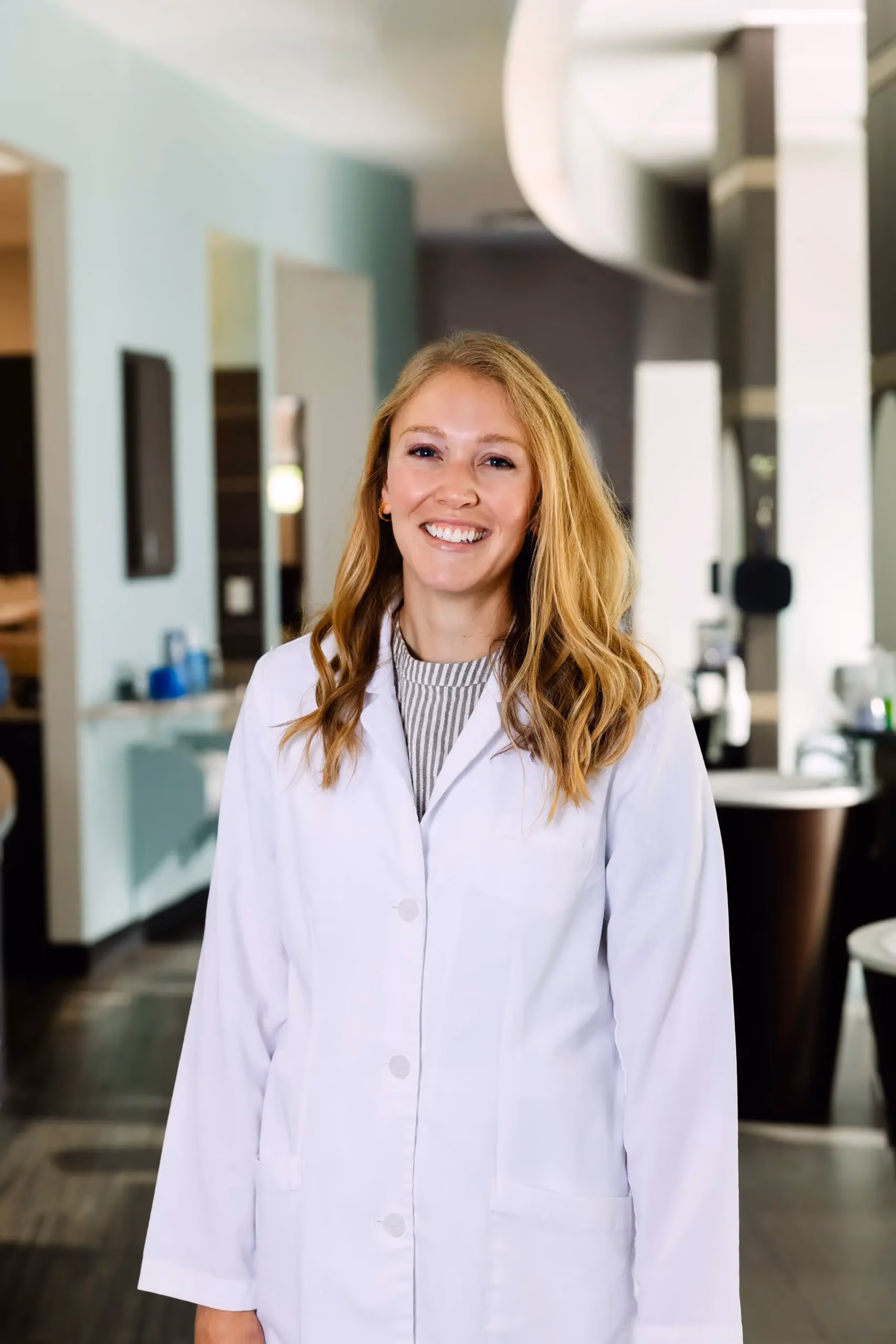 Smiling woman in a white lab coat standing in a modern medical or office setting.