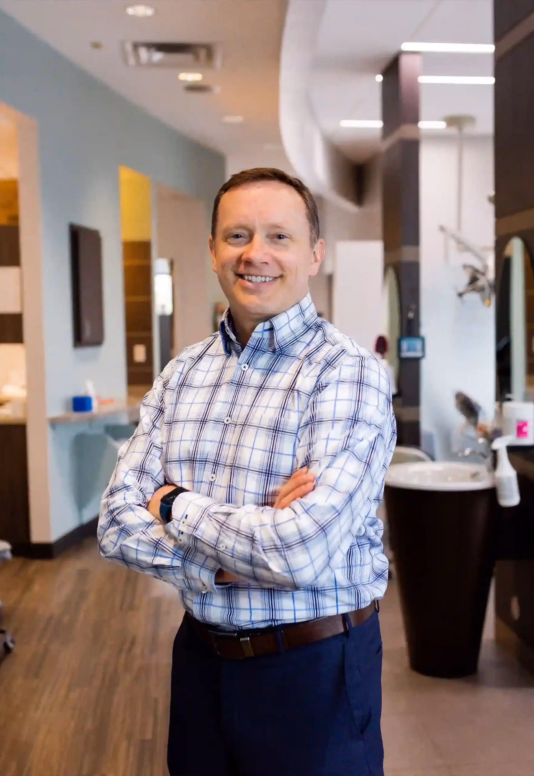 Smiling man with folded arms wearing a checkered shirt and dark pants in a modern office space.