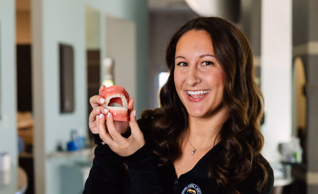 Smiling woman with long brown hair holding a dental model of teeth inside a room.