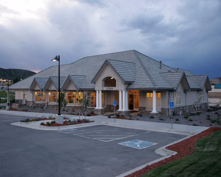 Large single-story building with grey tiled roof, stone facade, and lit windows at dusk with empty parking.