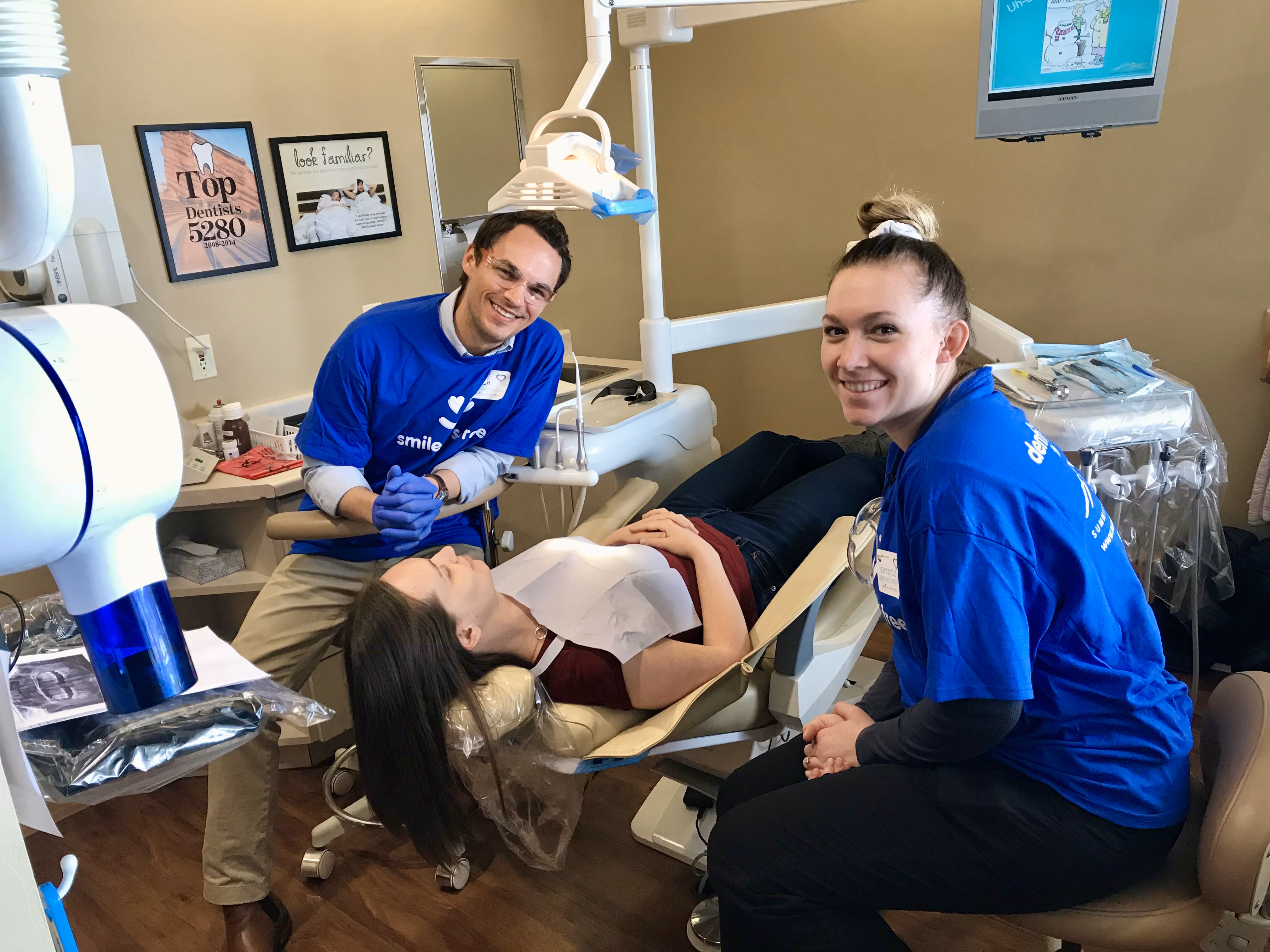 Dentist and assistant in blue shirts smiling with patient lying back in a dental chair.