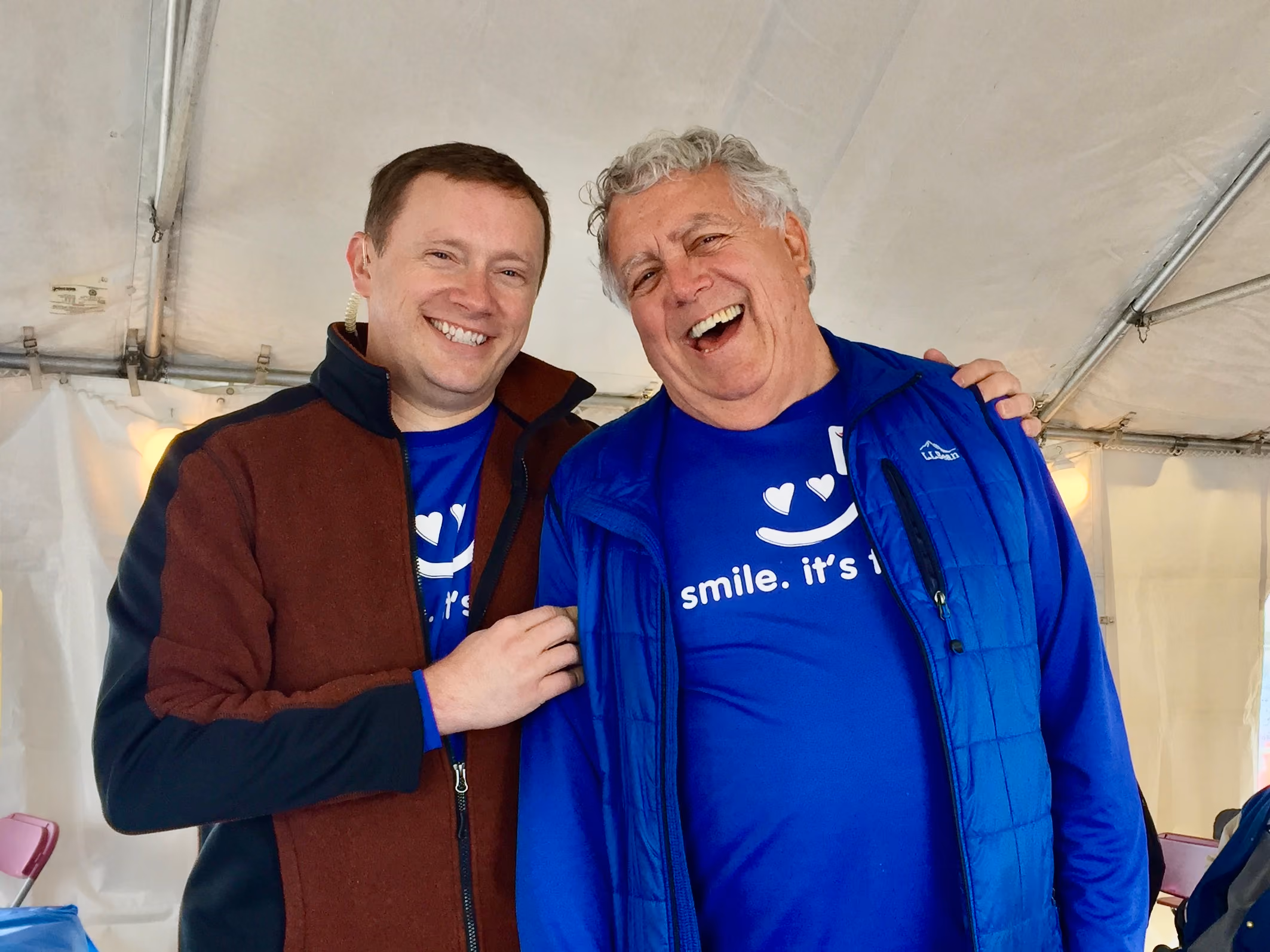 Two men smiling and posing together under a tent, one wearing a blue jacket and the other a brown one.