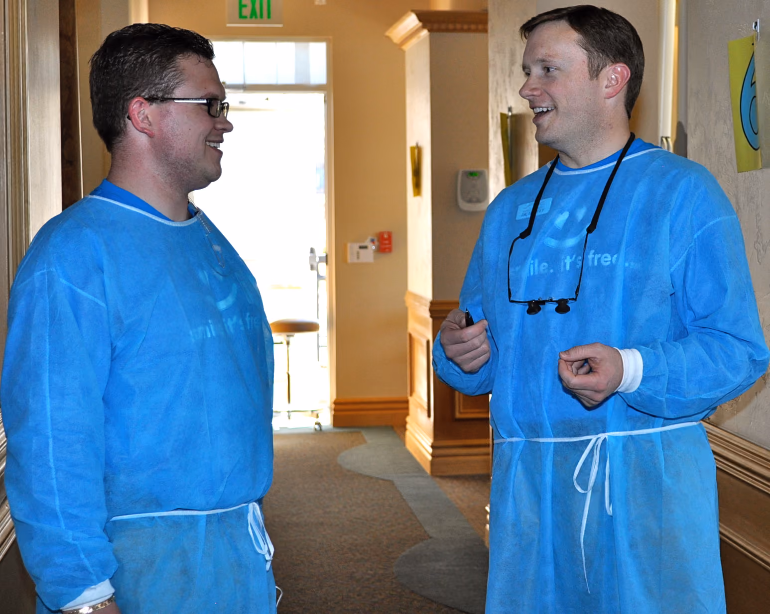 Two men wearing blue protective gowns talking and smiling indoors near an exit sign.