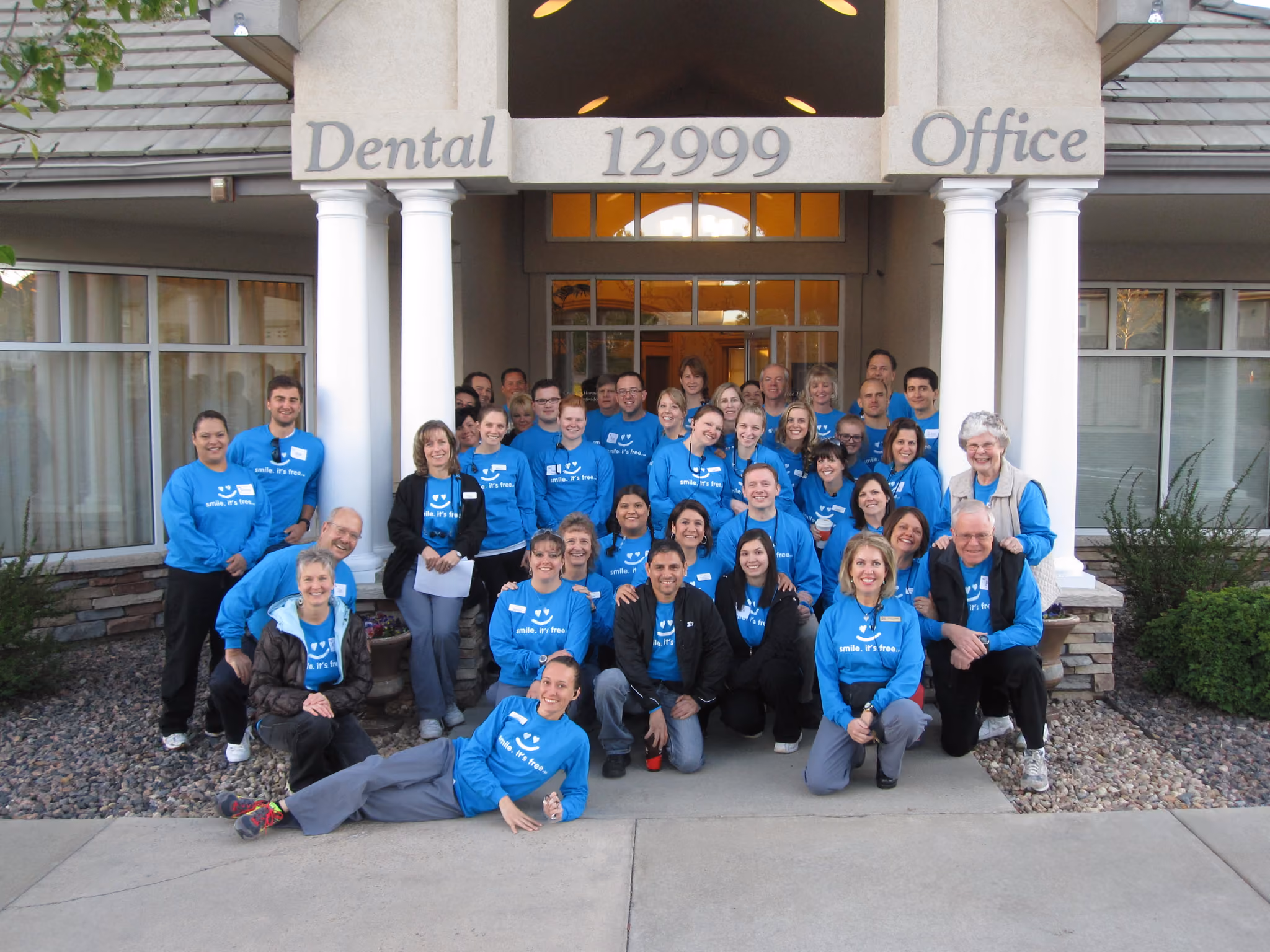 Group of people wearing blue shirts that say 'smile. It's free.' posing outside a dental office.