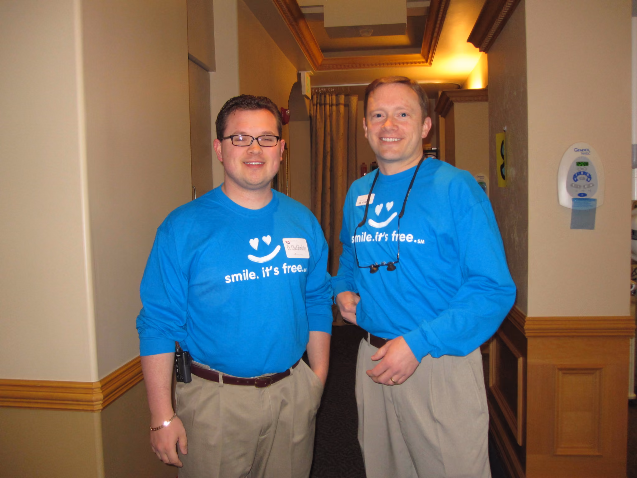 Two men in blue shirts with 'smile. it's free.' text standing in a hallway, smiling at the camera.