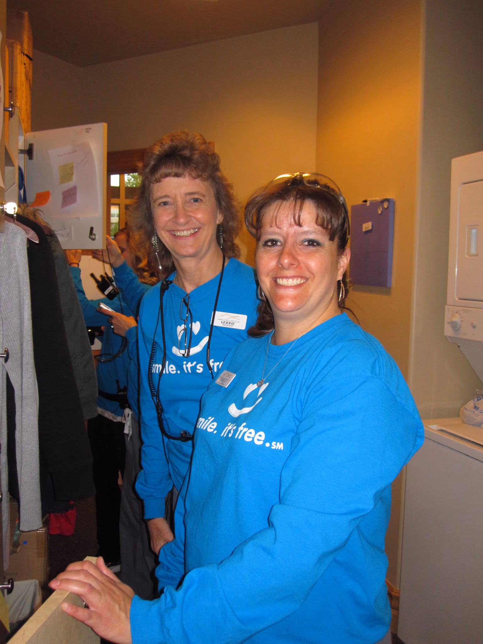 Two smiling women in blue shirts reading 'smile. it's free.' indoors with coats and a washer nearby.