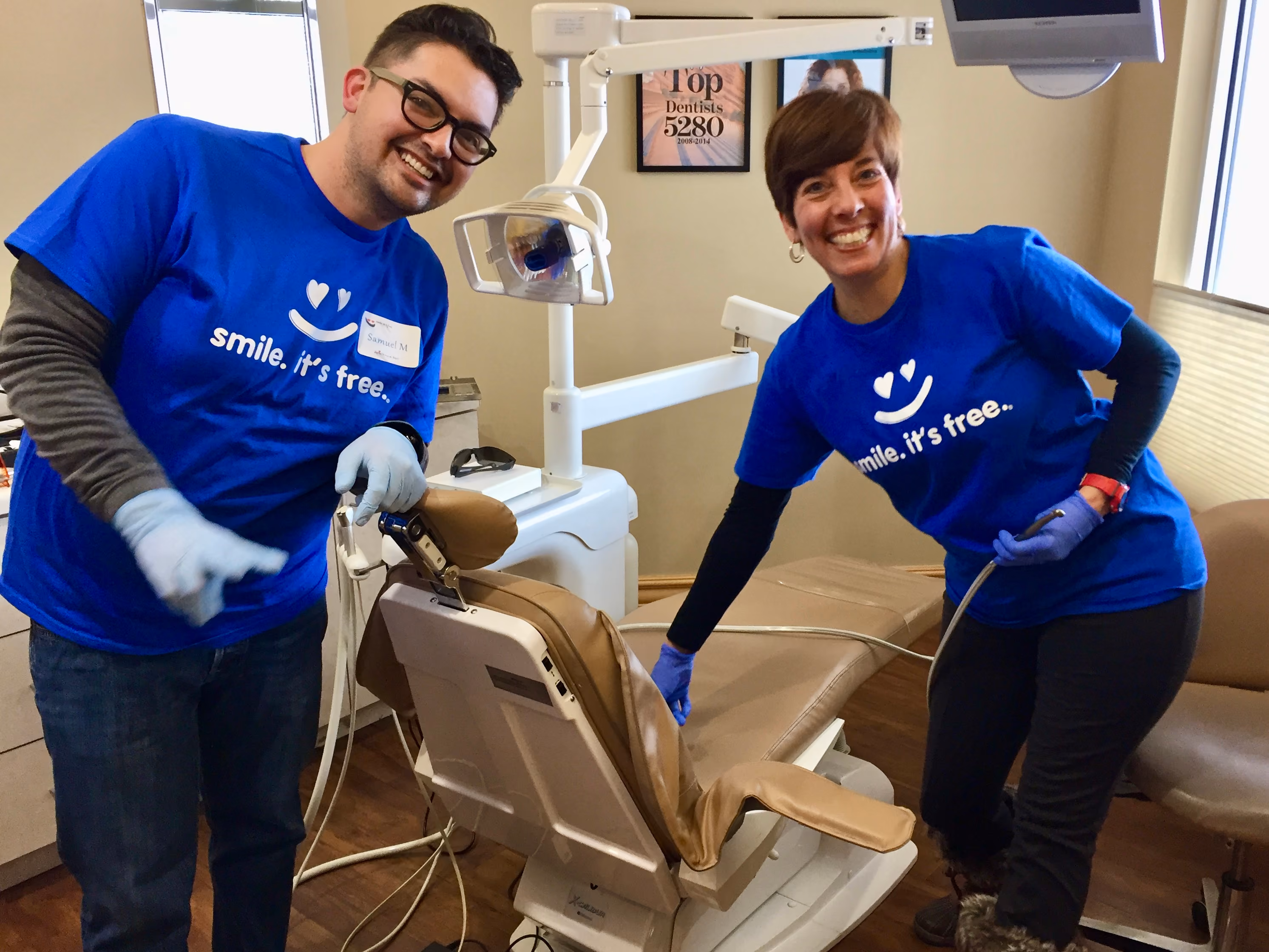 Two people in blue shirts with 'smile. it's free.' smiling beside a dental chair in a clinic.