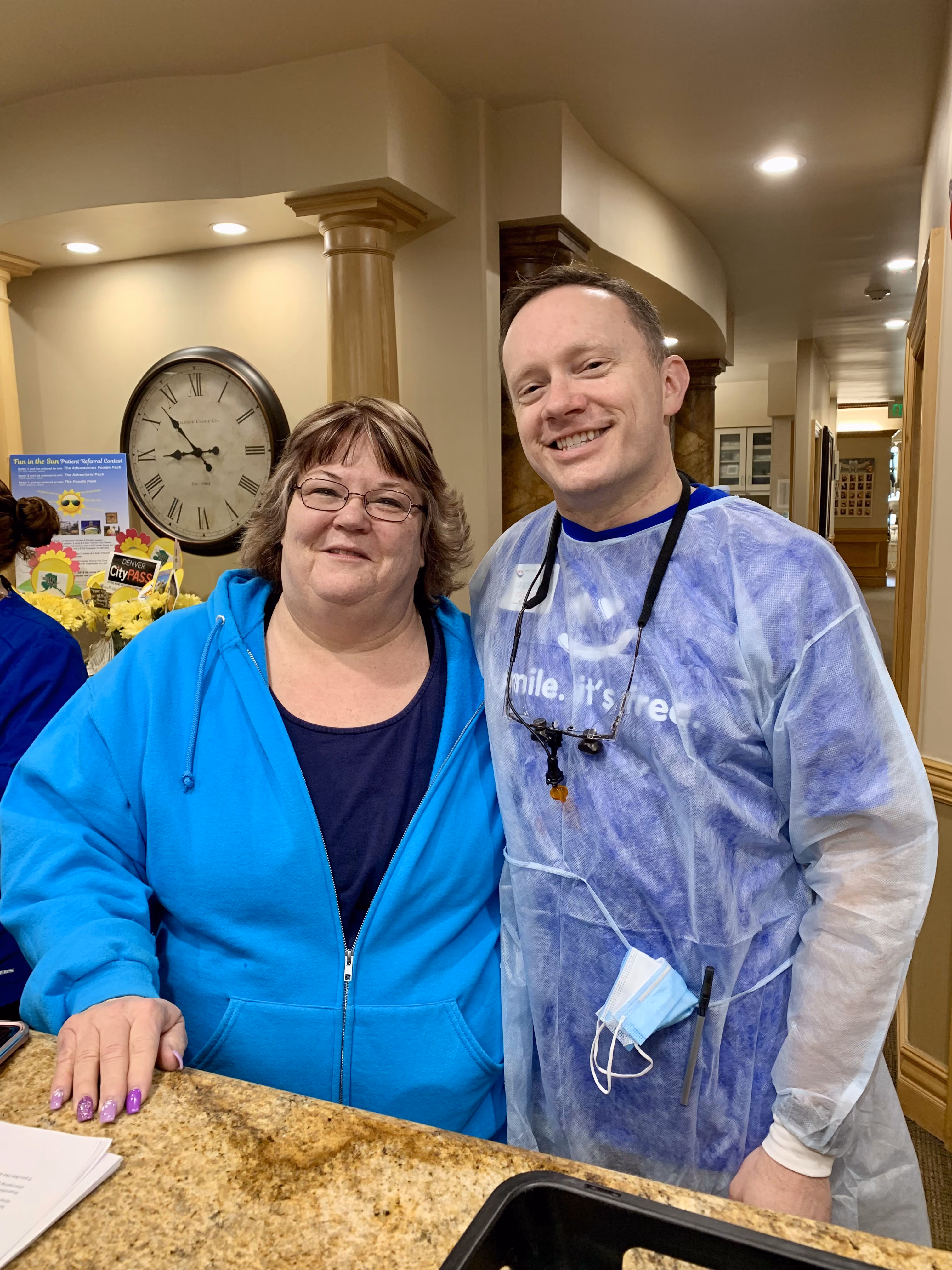 Smiling woman in blue hoodie and man in a blue gown standing together at a counter indoors.