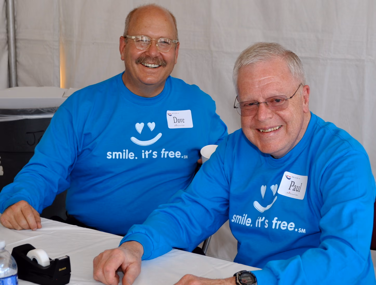 Two smiling men wearing blue shirts with 'smile. it's free.' sitting at a table with name tags.