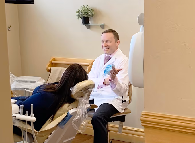 Dentist in white coat discussing dental care with a patient reclined in a dental chair.