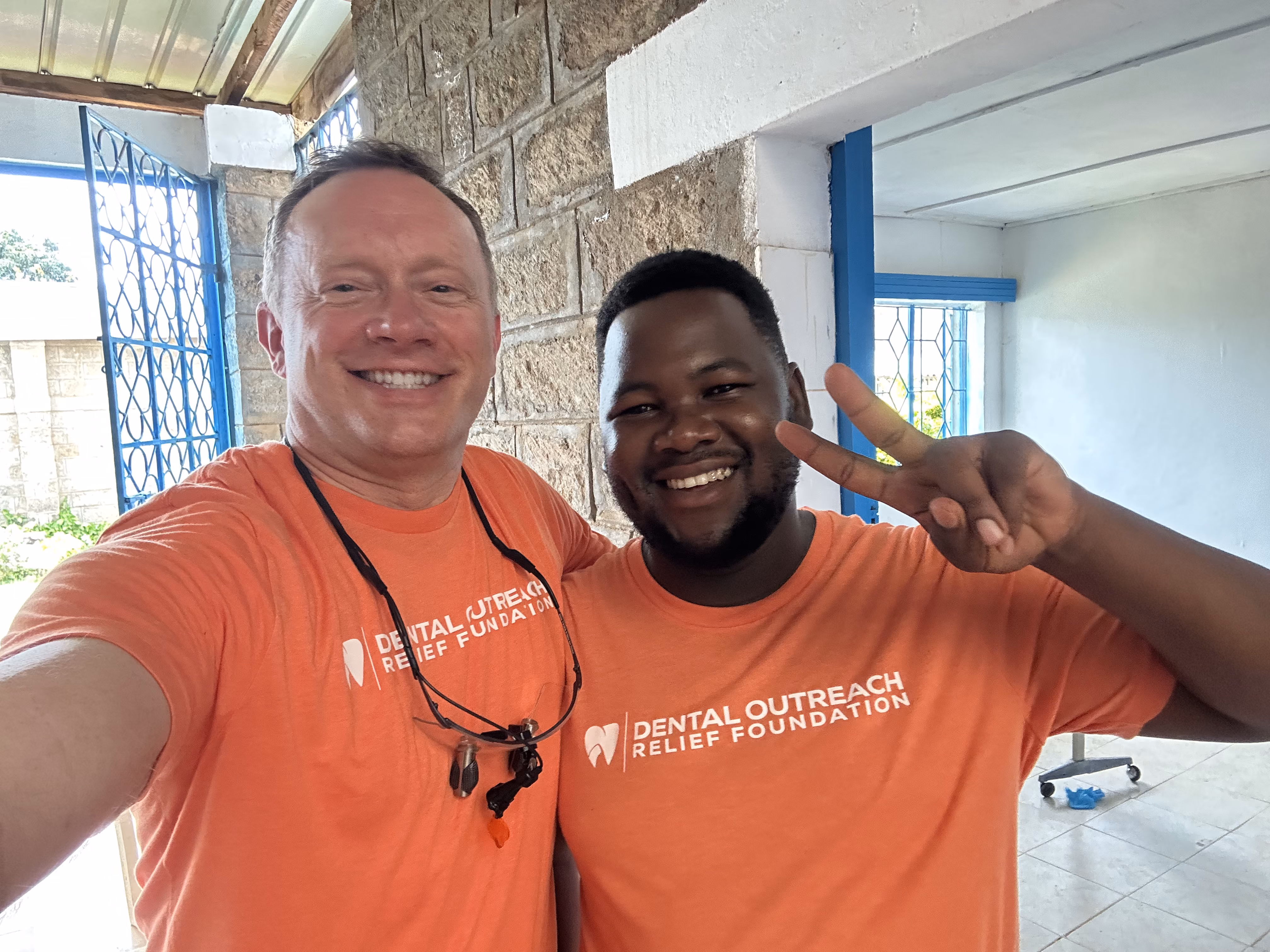 Two men in orange Dental Outreach Relief Foundation shirts smiling, one flashing a peace sign.