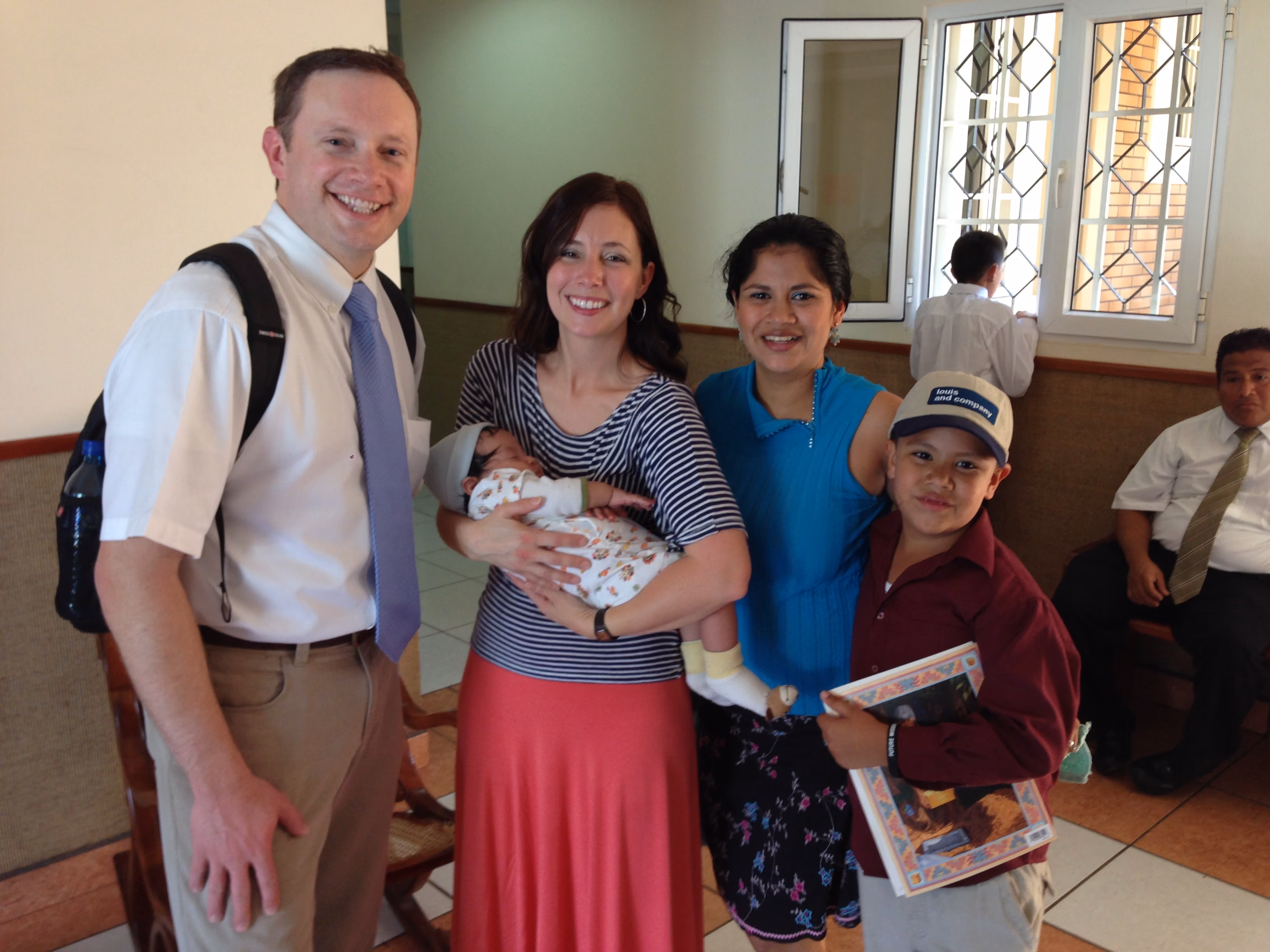Group of five smiling people inside a room, including a man, two women, a boy with a book, and a baby.