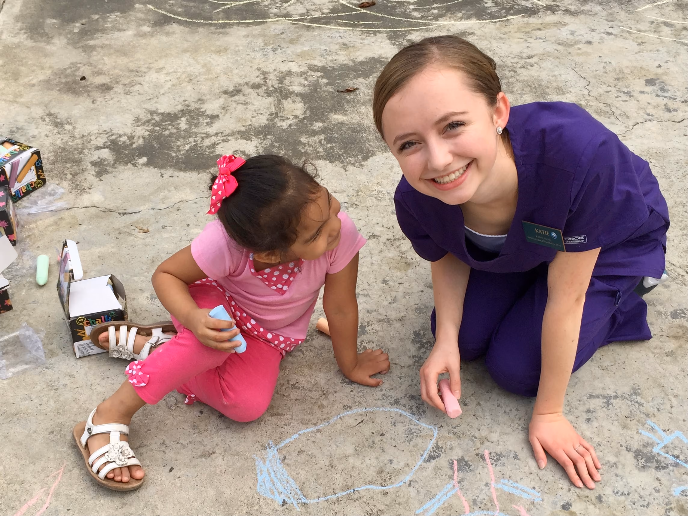 Young girl and woman in purple scrubs drawing with sidewalk chalk and smiling outdoors.