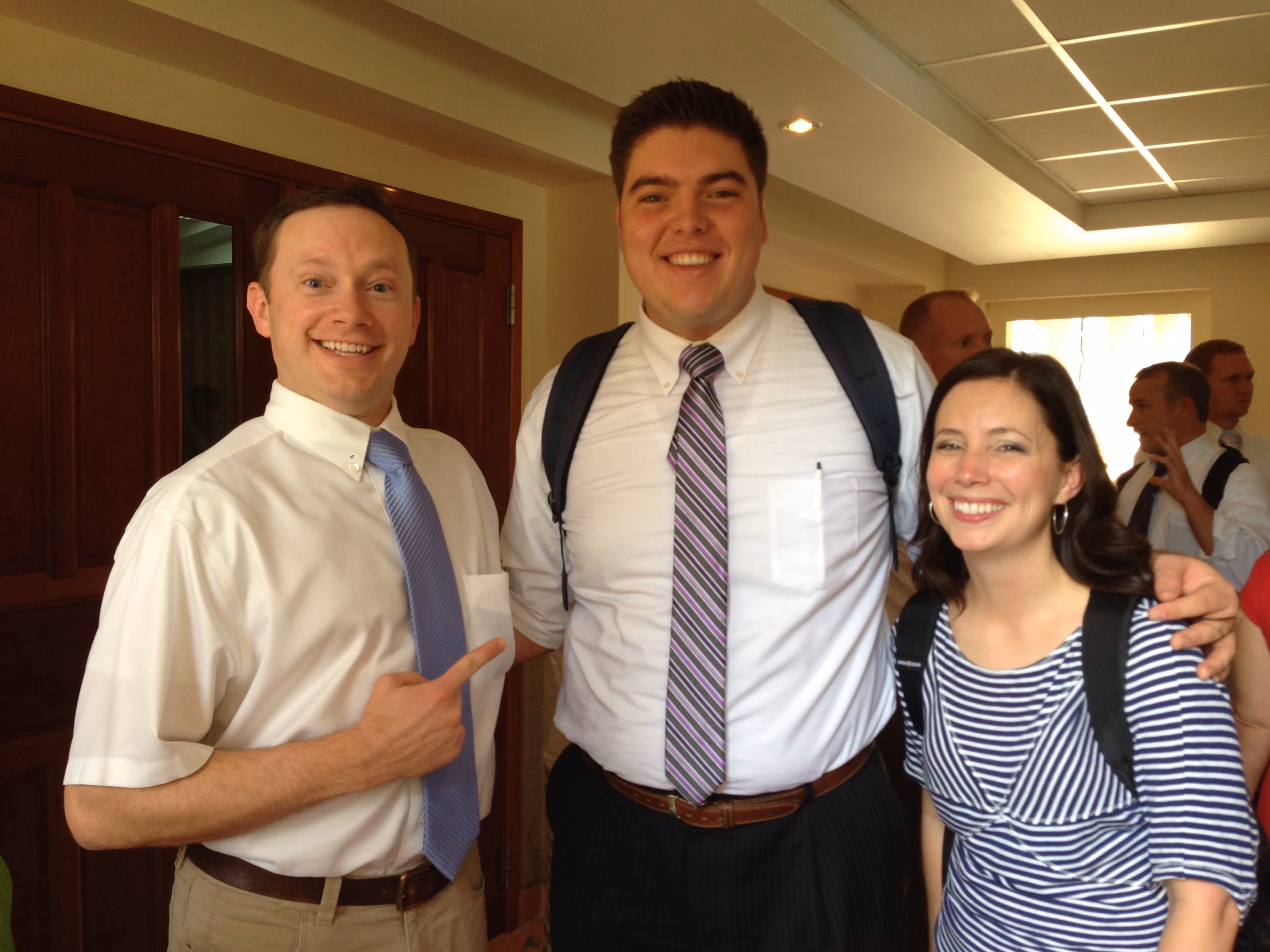 Three smiling adults posing indoors, two men in dress shirts and ties, one woman in a striped top.