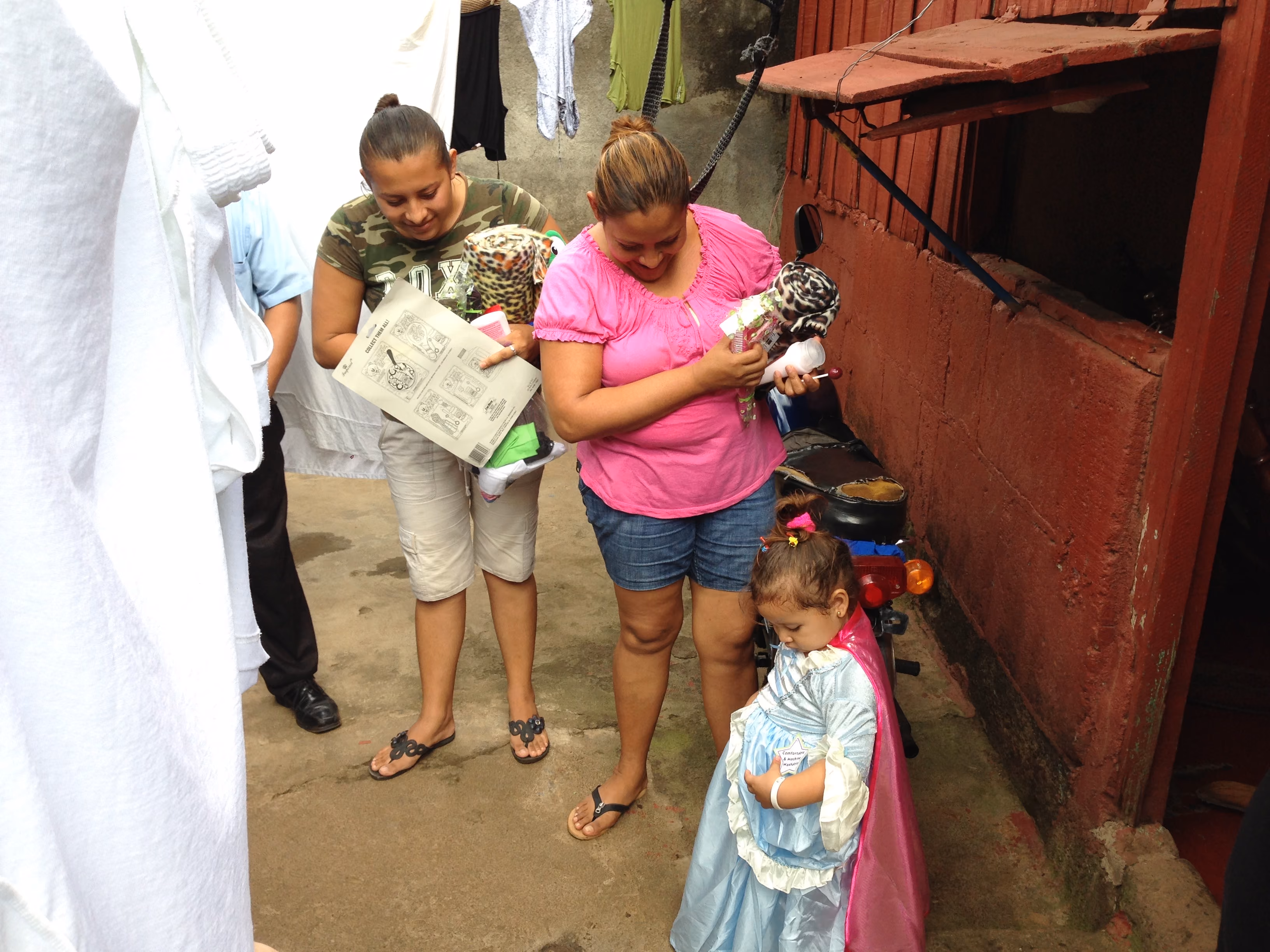 Two women with toys and a young girl dressed as a princess standing in a narrow outdoor corridor.