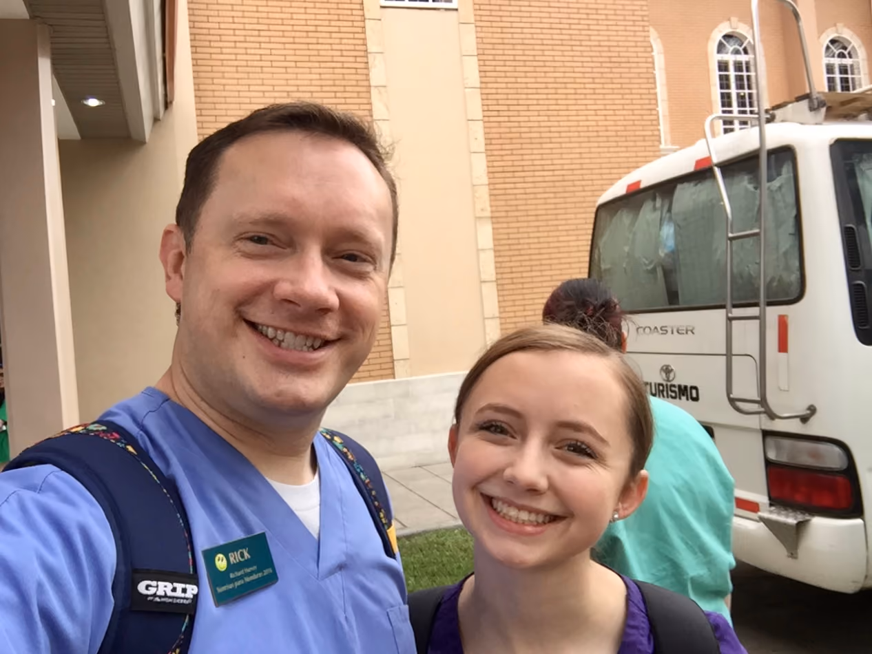 Two smiling healthcare workers in scrubs with backpacks standing outside near a bus and brick building.
