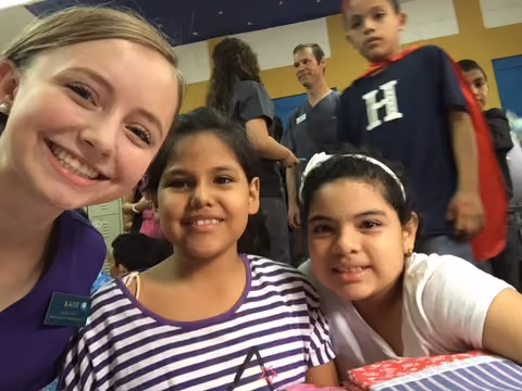 A smiling woman with two young girls in a striped and white shirt, posing indoors with other people in background.