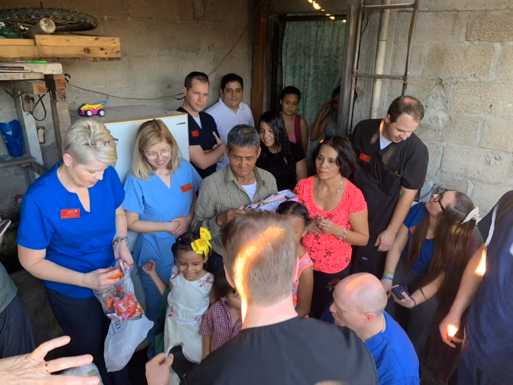 Group of adults and children interacting in a modest room, some wearing medical scrubs and name tags.