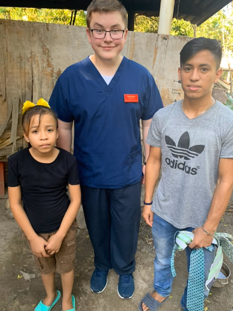 Three people standing outside: a young girl with a yellow bow, a man in navy scrubs, and a young man holding fabric.