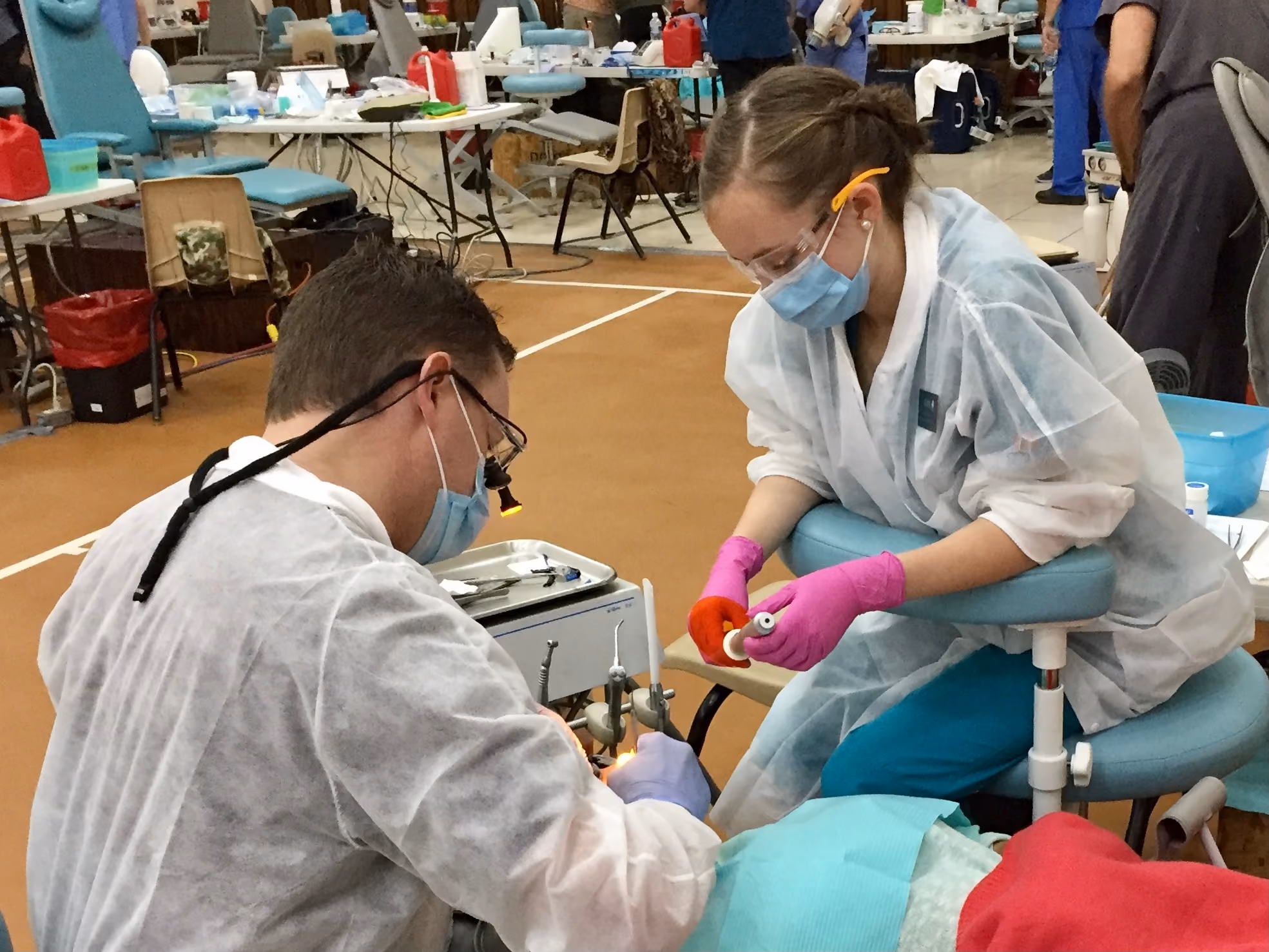Dentist treating a patient while an assistant prepares dental tools in a clinic setting.