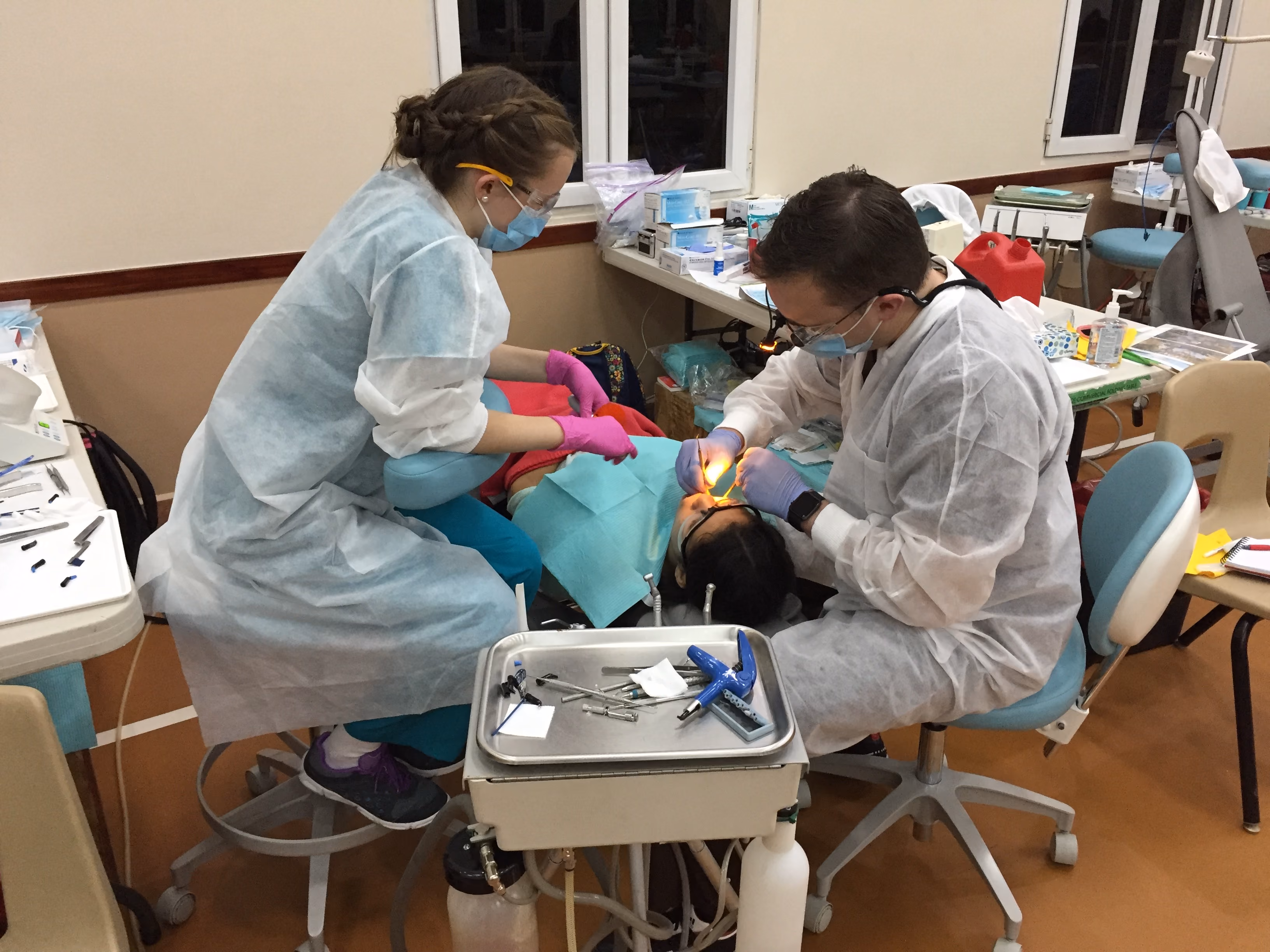 Two dental professionals in protective gear treating a patient lying on a chair in a clinic.