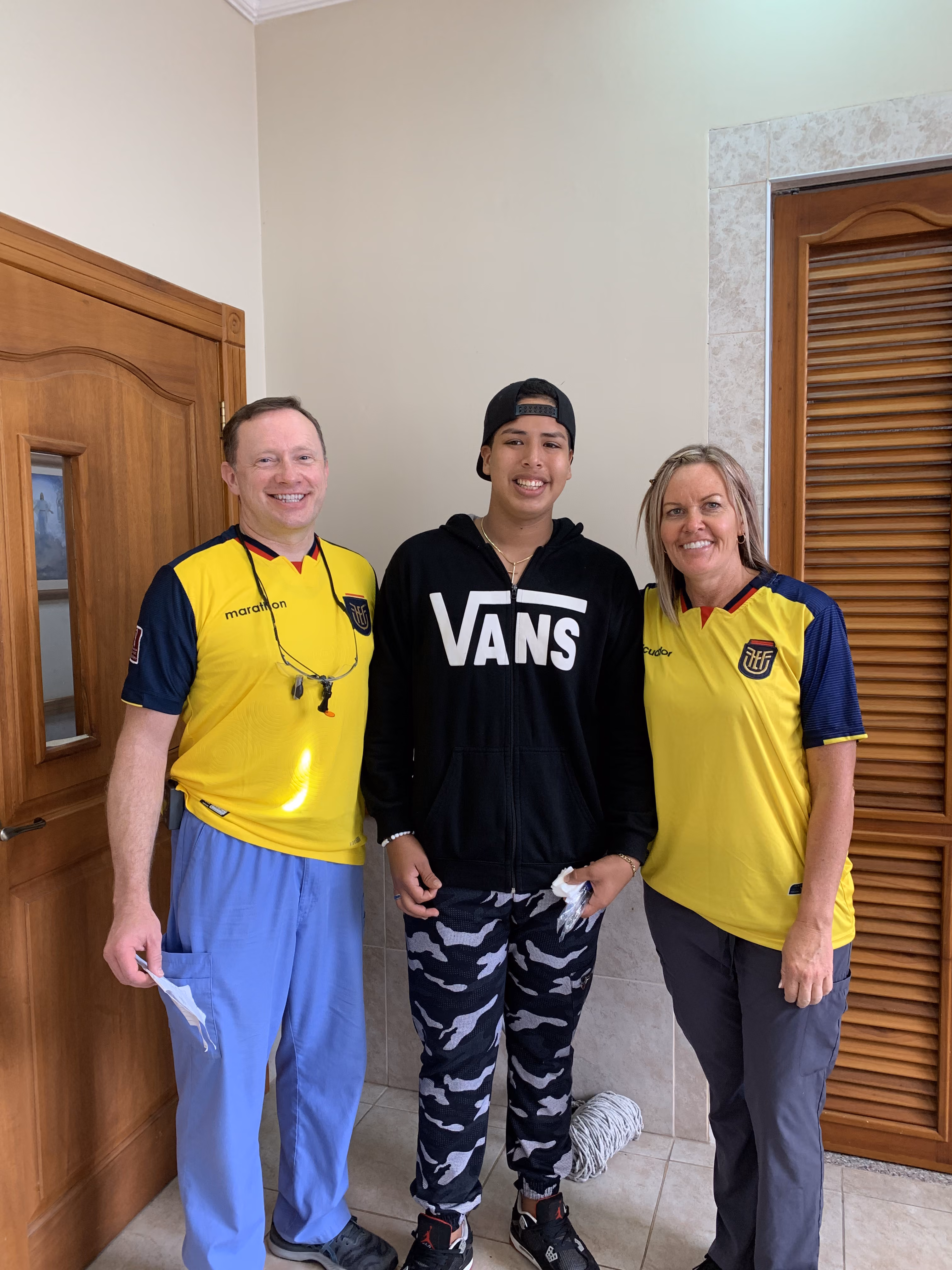 Three people standing indoors: two in yellow Ecuador soccer jerseys and one in a black Vans hoodie.