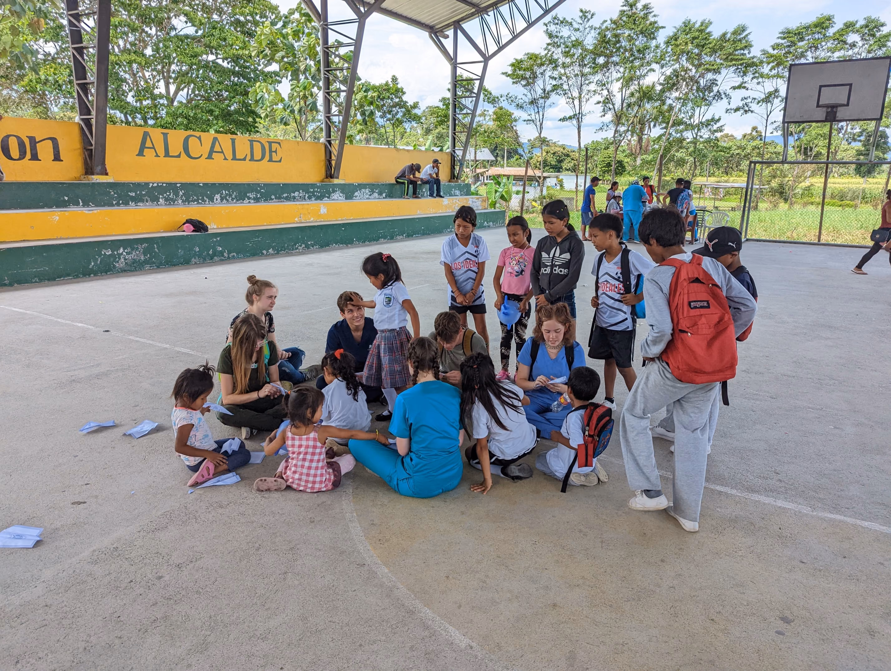Group of children and adults gathered in a covered outdoor court with yellow and green bleachers.