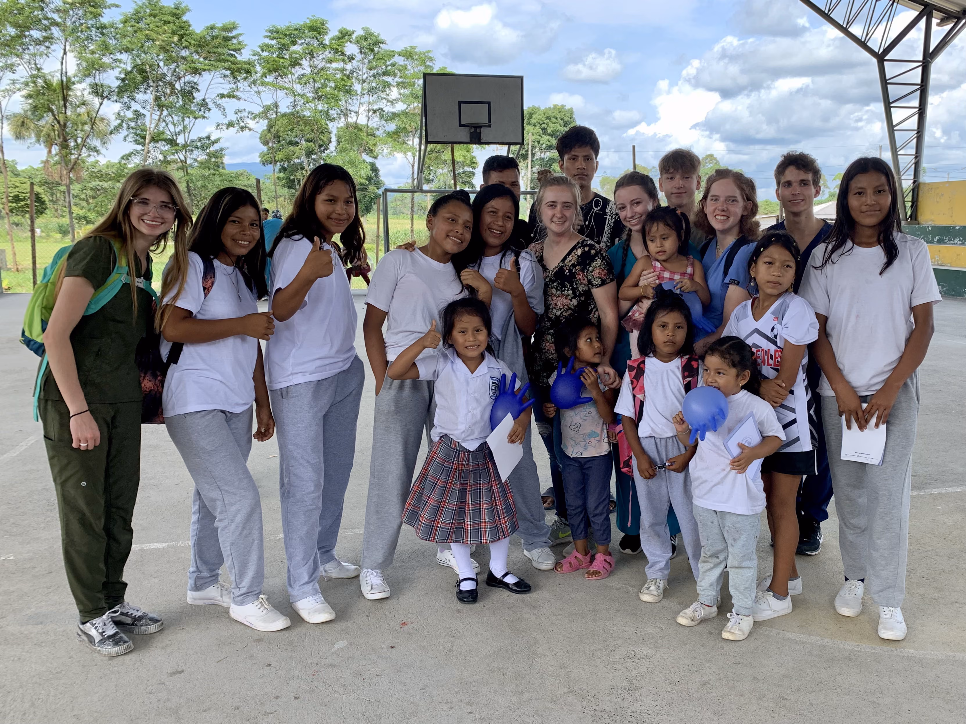 Group of smiling children and young adults posing together under an outdoor basketball court roof.