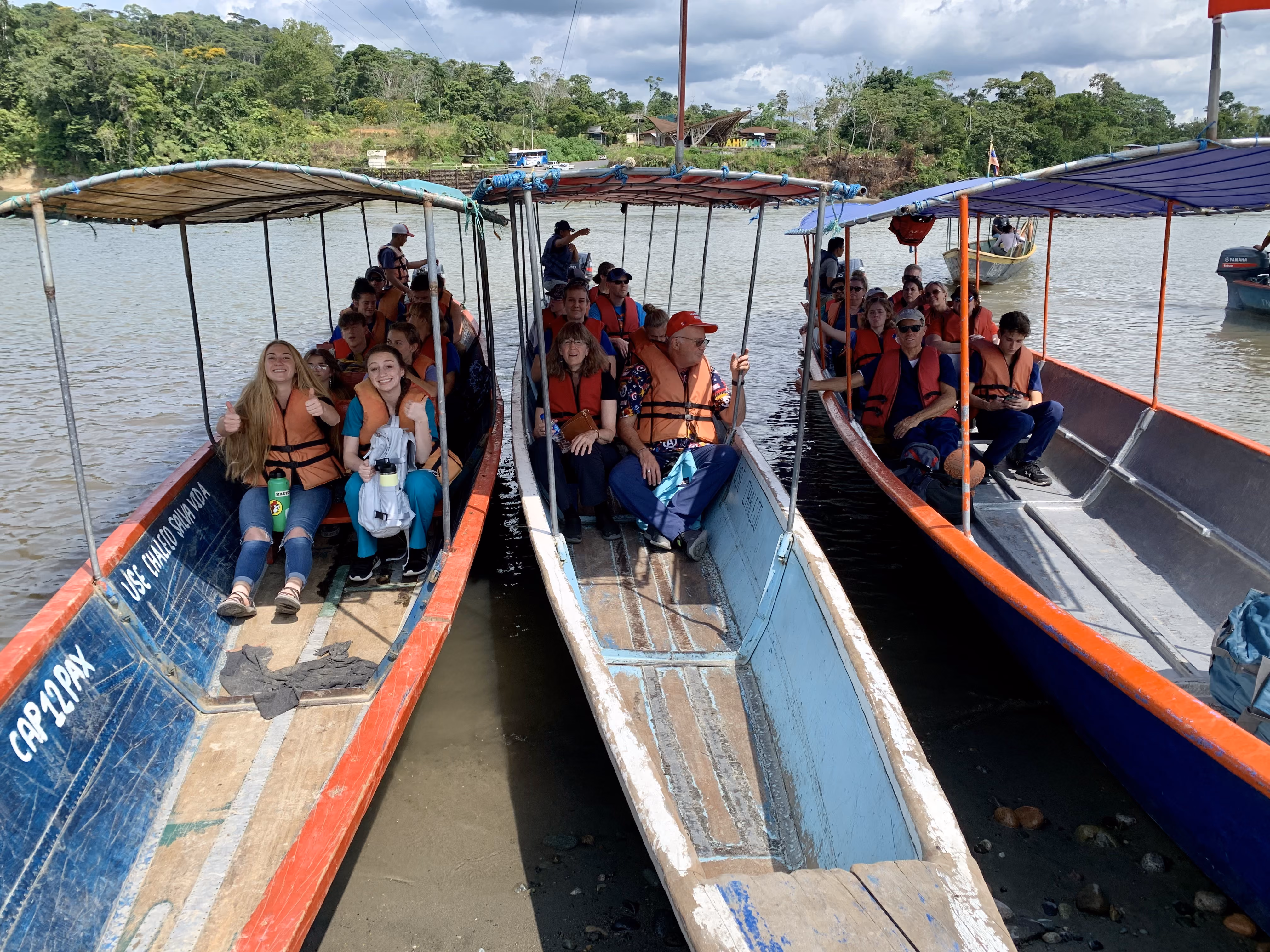 Groups of people wearing life jackets sitting in three covered boats docked on a river near a forested shore.