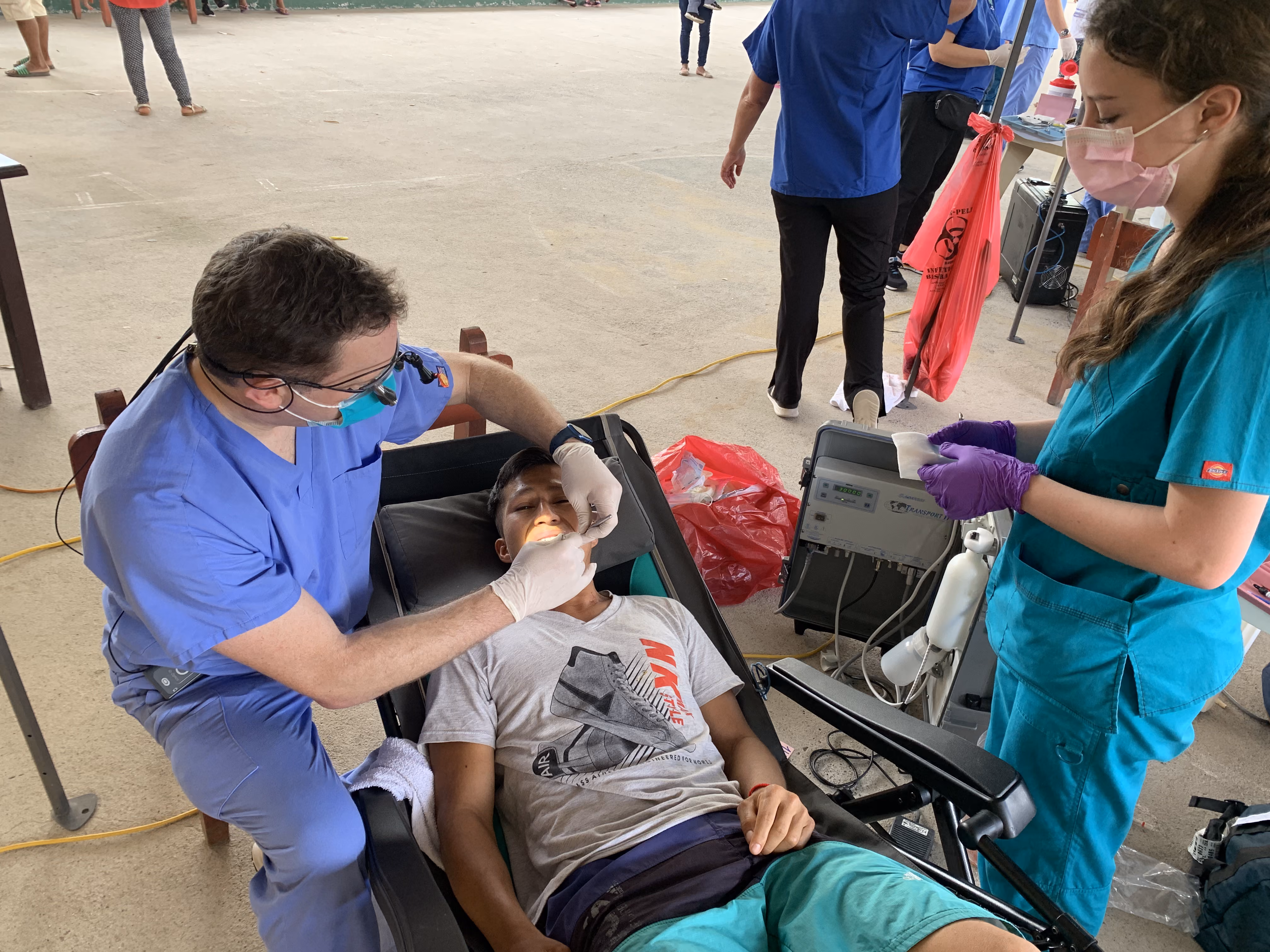Medical professional in blue scrubs treating a young man reclining in a chair with assistance nearby.