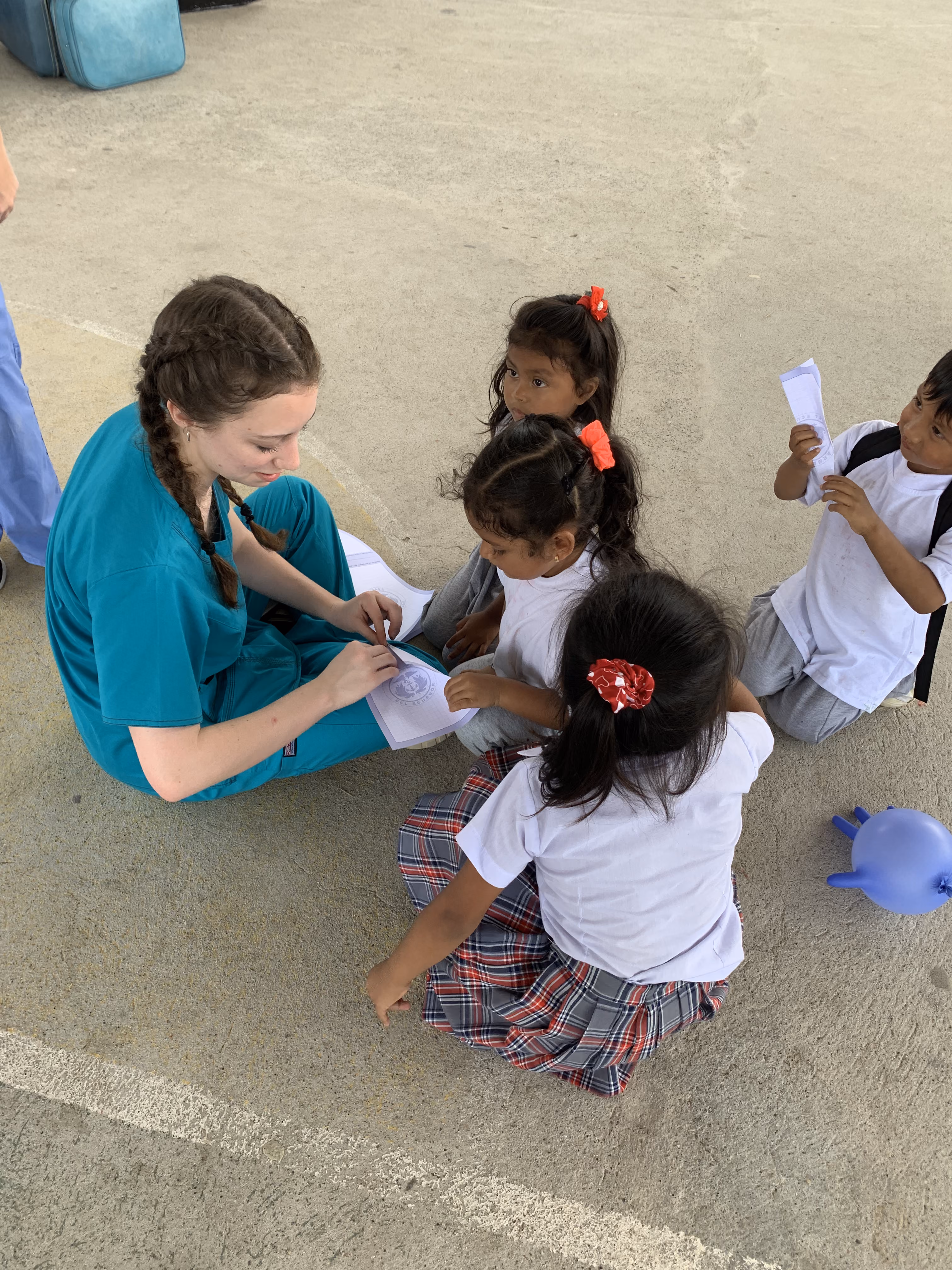 Woman in teal scrubs sitting on ground with four children holding papers and a balloon nearby.