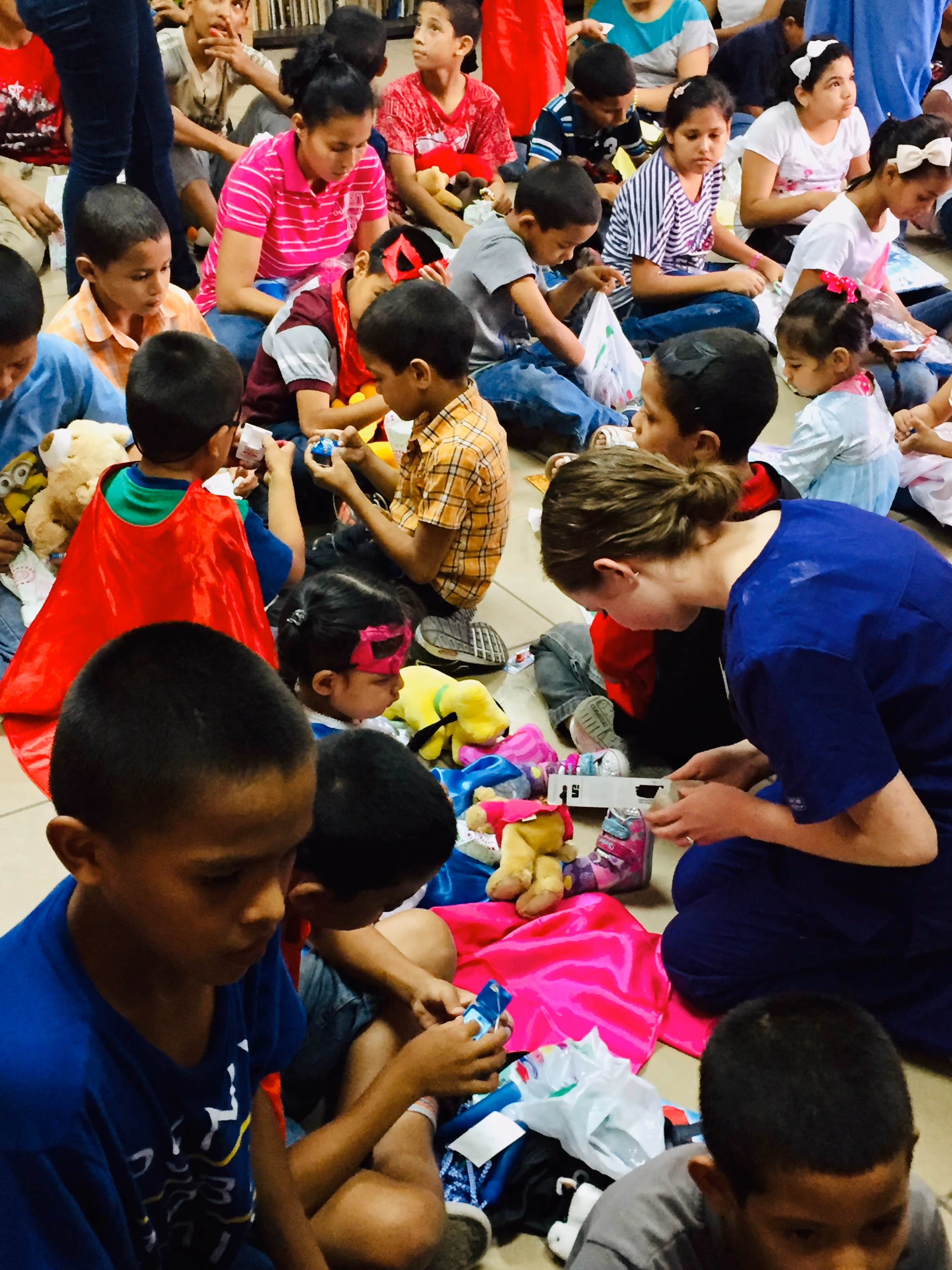 Group of children and a woman sitting on floor, opening toys and gifts in a community setting.