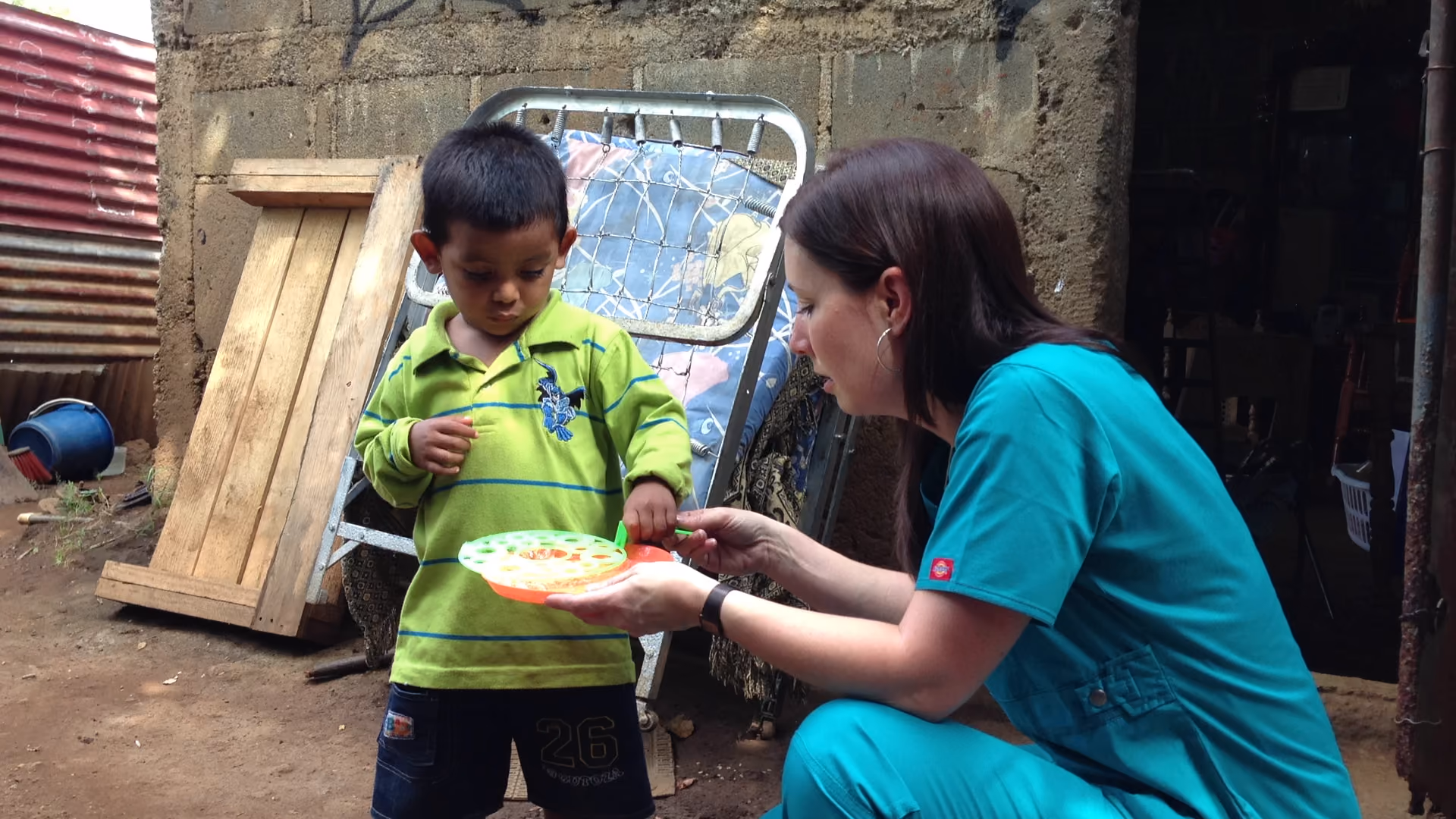 Woman in teal scrubs helps a young boy with a toy or game inside a rustic outdoor area.