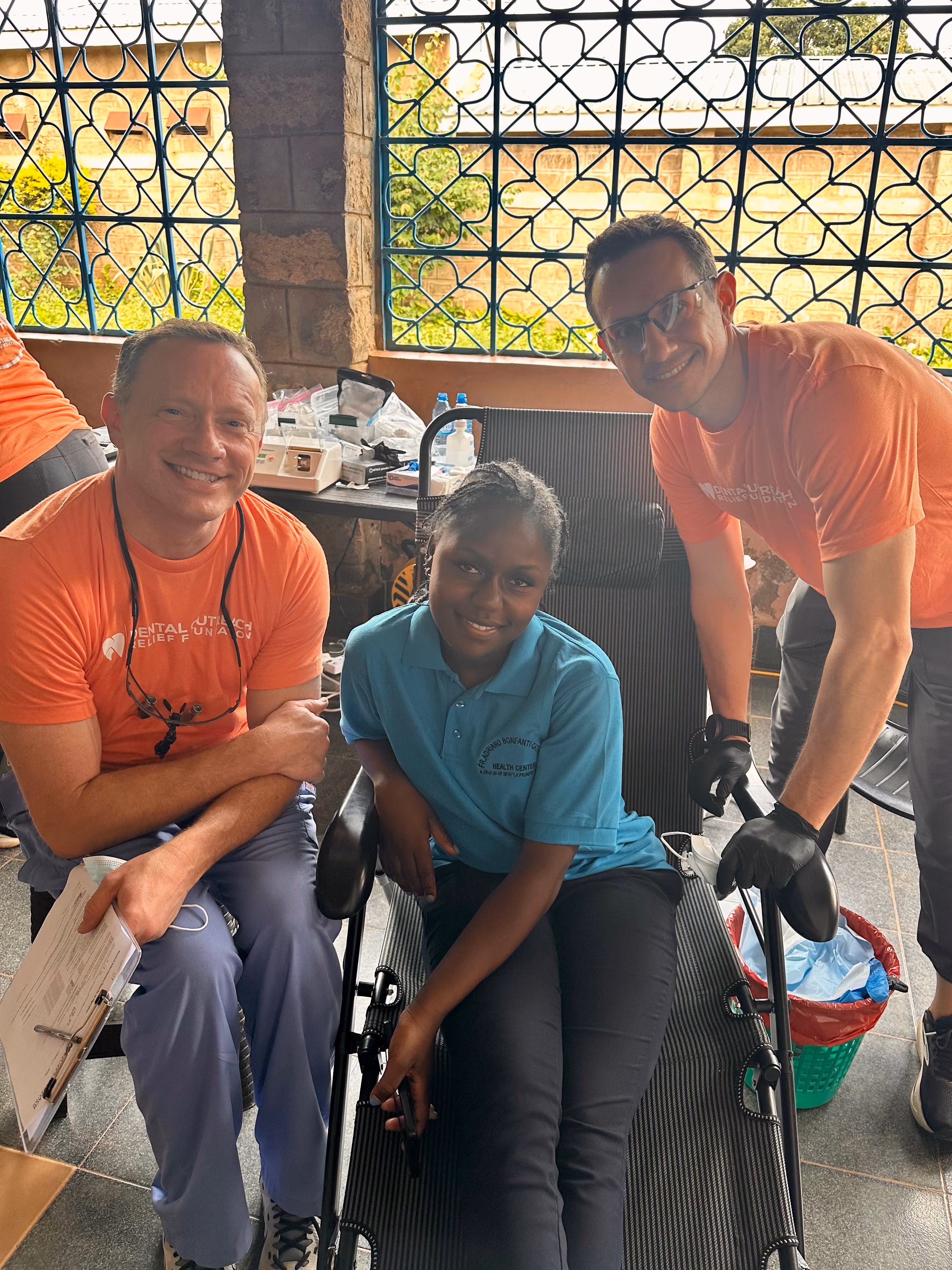 Two men in orange shirts smile beside a woman reclining on a chair in a room with decorative windows.