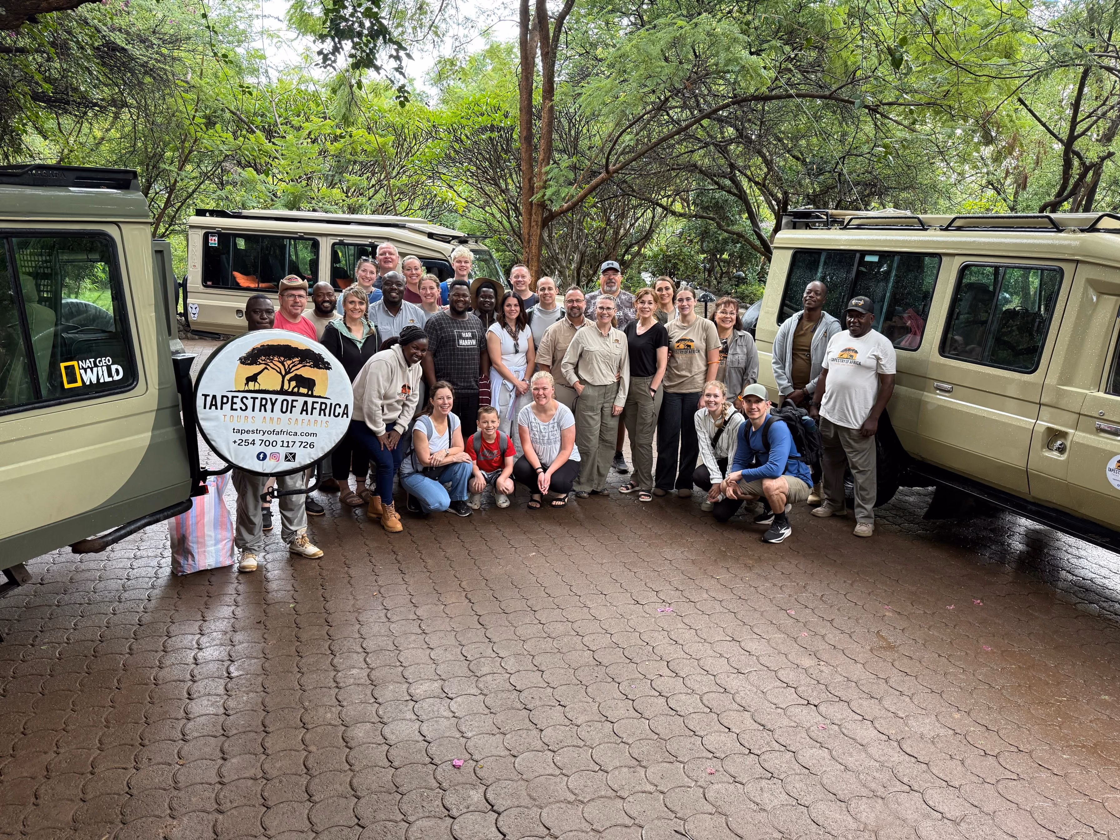 Group of smiling safari tourists posing with guides between two safari vehicles under trees.