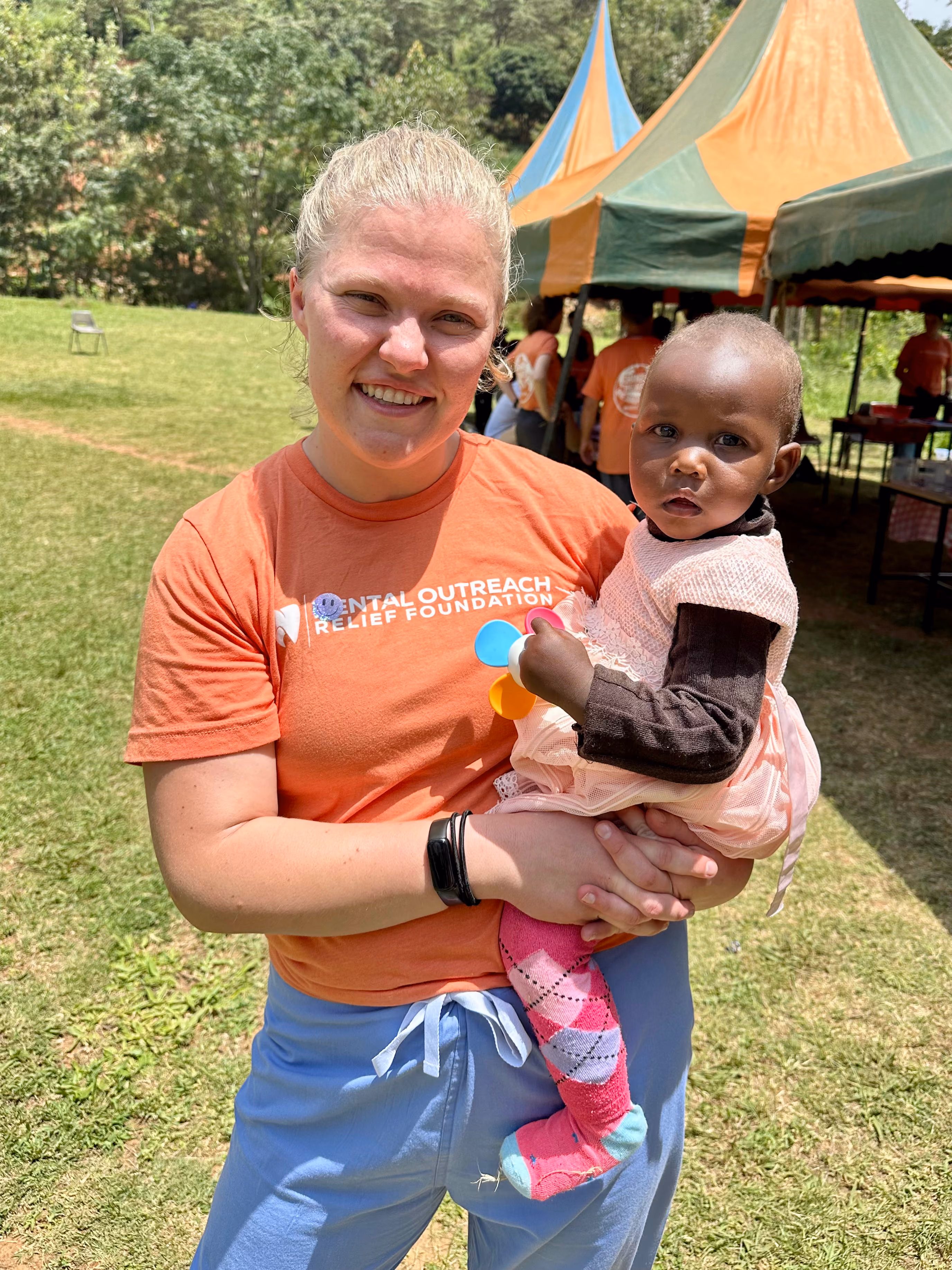 Volunteers in orange shirts interacting and handing out materials to school children in uniforms outdoors.