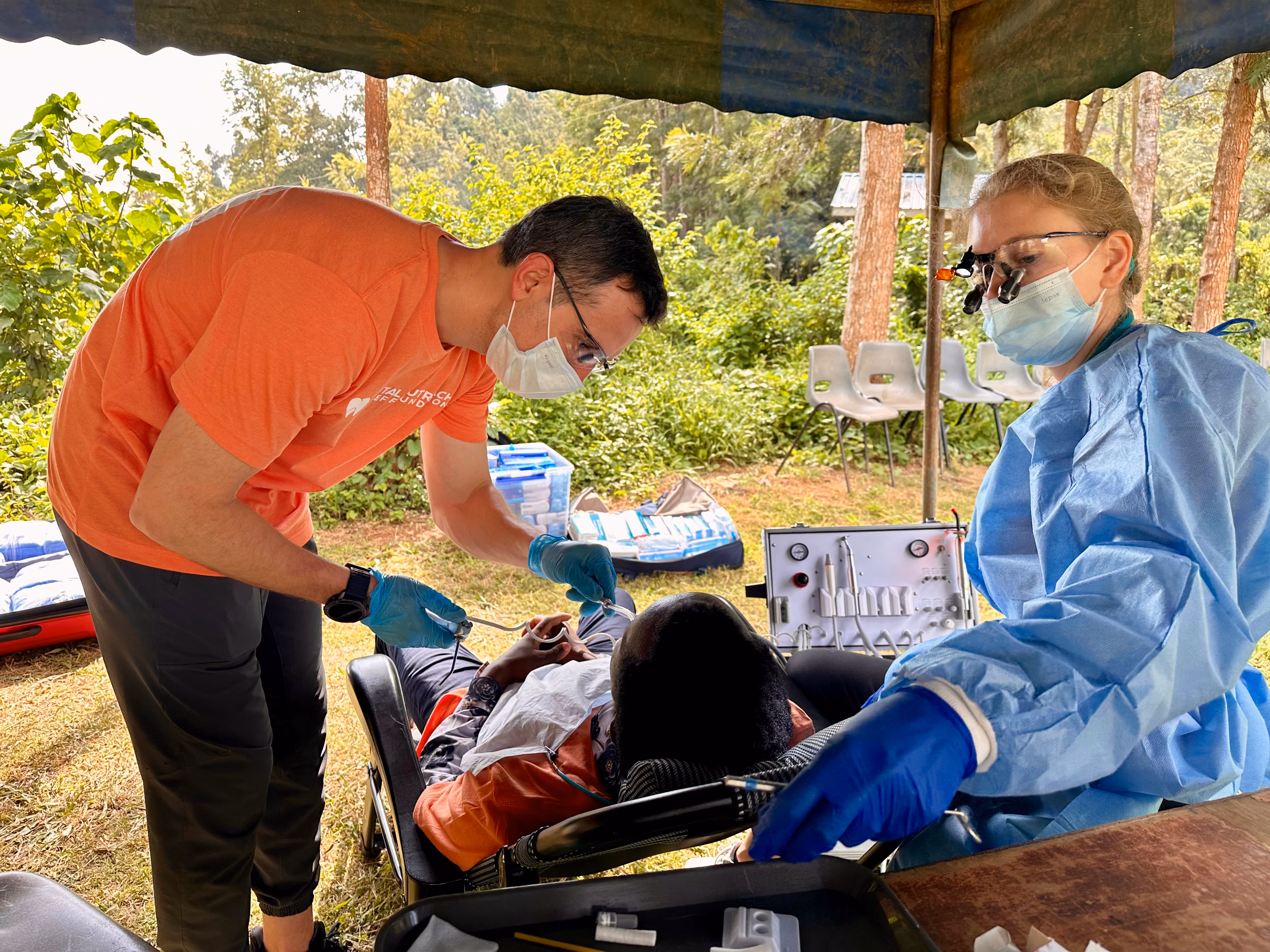 Healthcare workers in protective gowns treating a patient outdoors under a tent with medical supplies nearby.