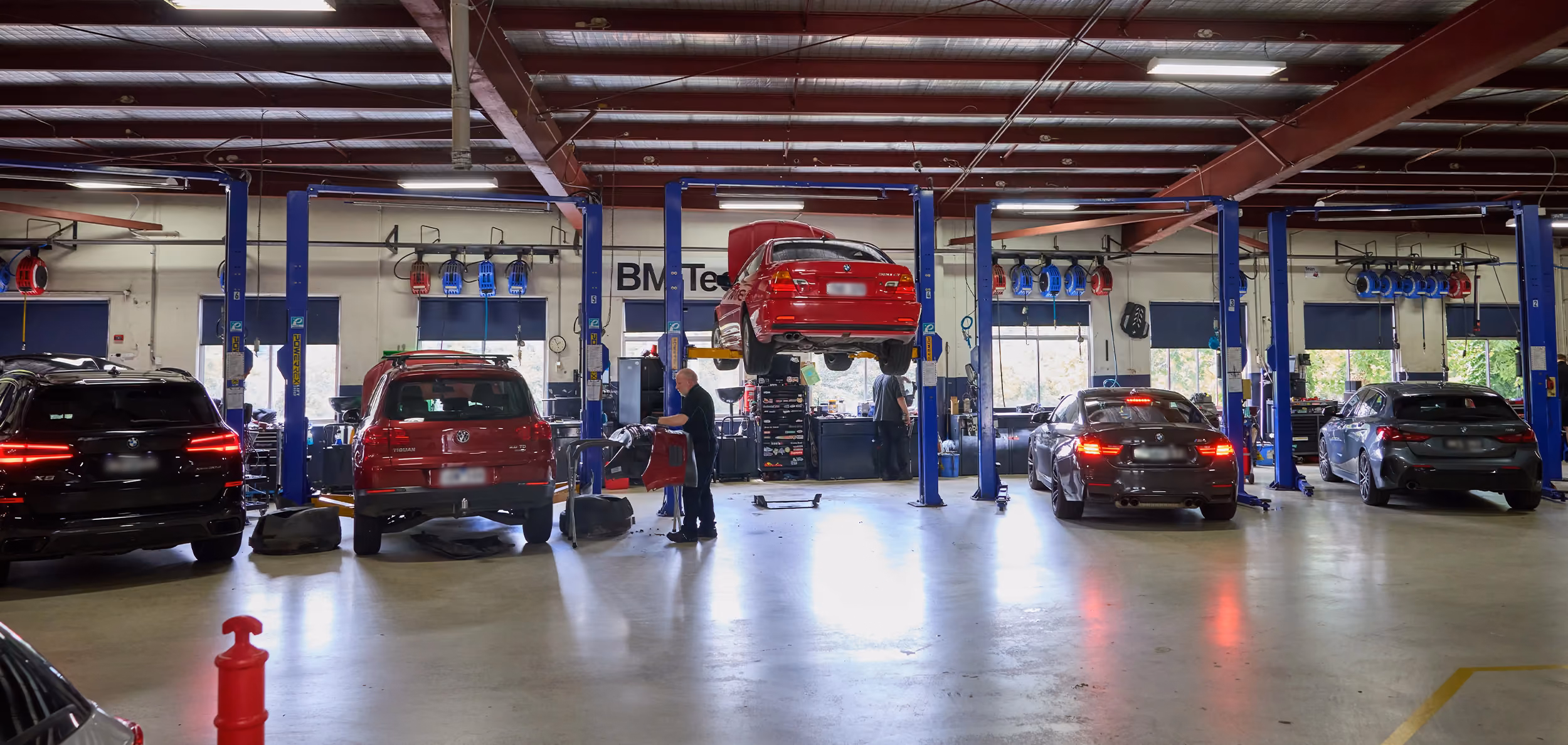 Interior of a spacious auto repair workshop with multiple blue car lifts, various cars parked and being serviced, and two mechanics working under a red car elevated on a lift.