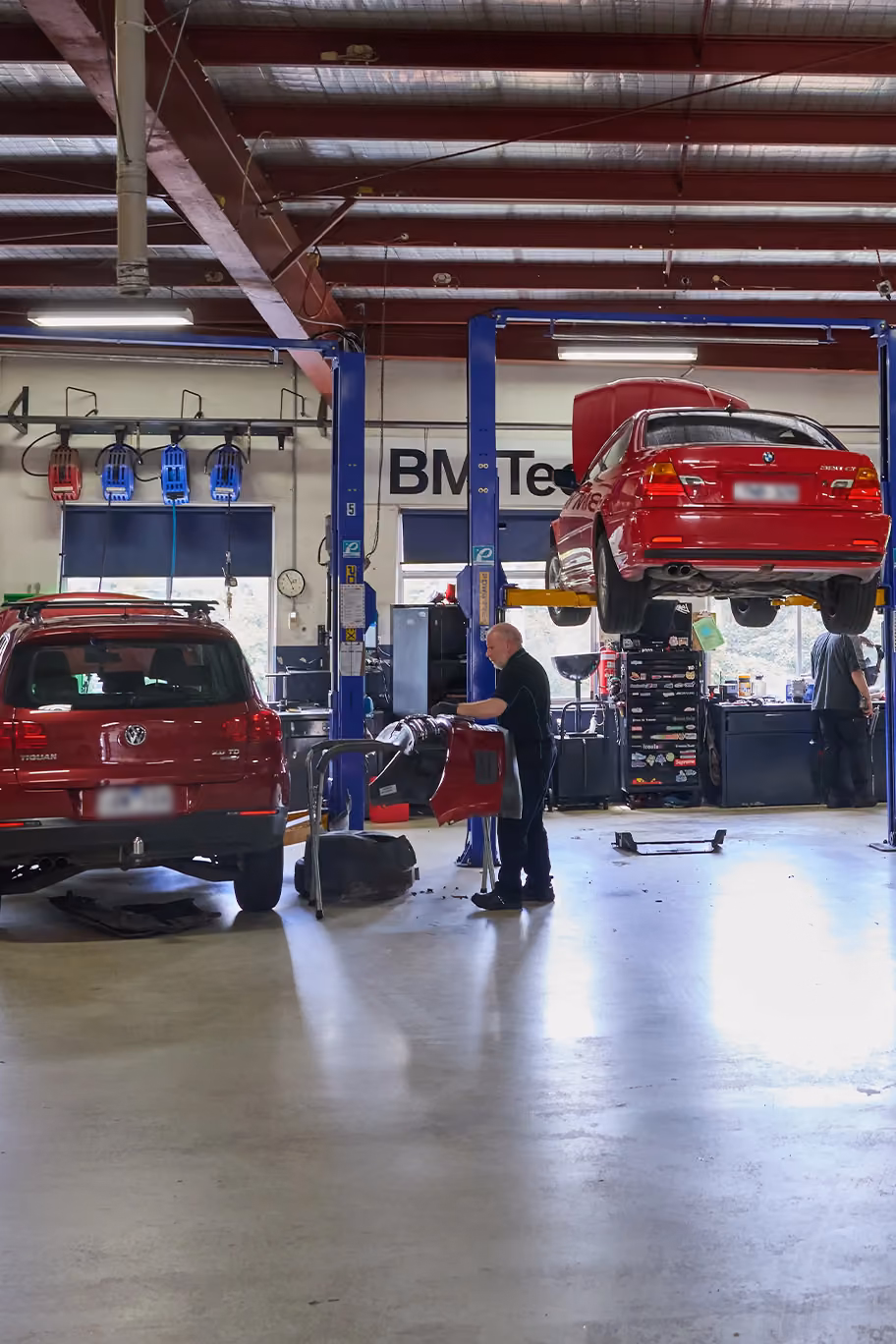 Interior view of a car workshop with two red cars, one on the ground and one lifted on a blue hydraulic lift. Two mechanics work on the cars under a metal ceiling with exposed beams and fluorescent lights. Tools and equipment are visible along the walls.