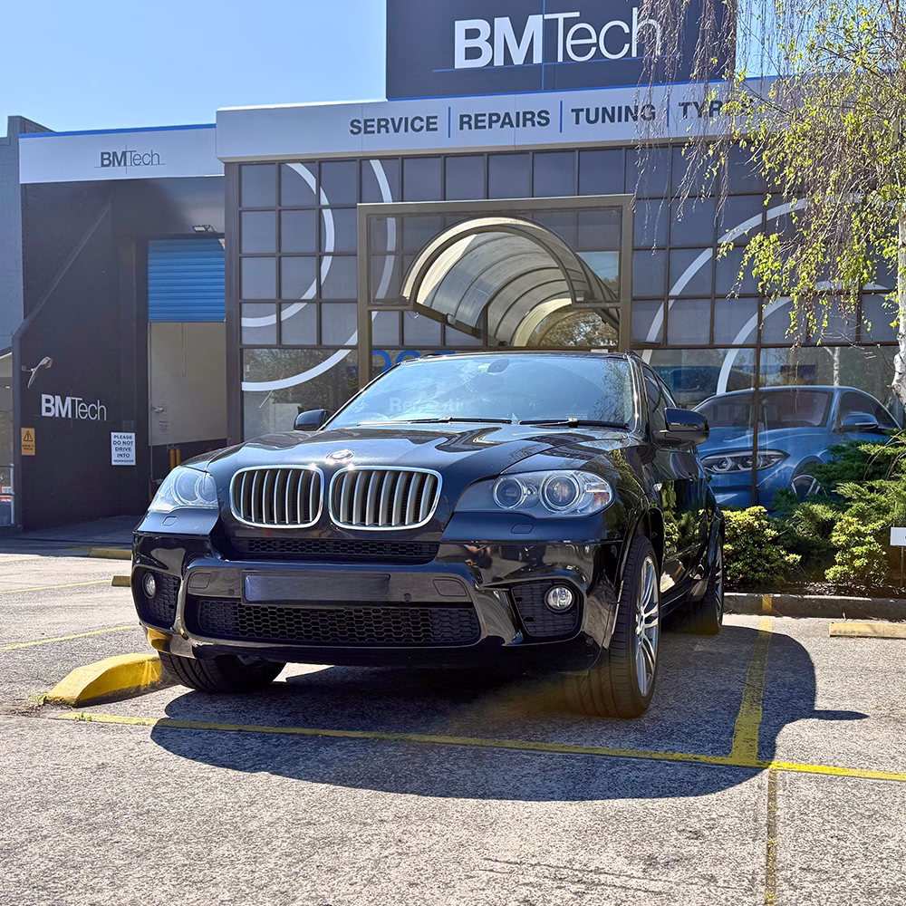 A mechanic near a green machine, next to a white BMW SUV on a lift with its hood up.