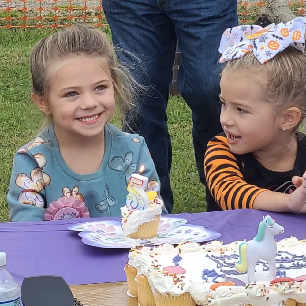 Two young girls at a birthday party with cupcakes, one smiling next to a lit candle number 5.