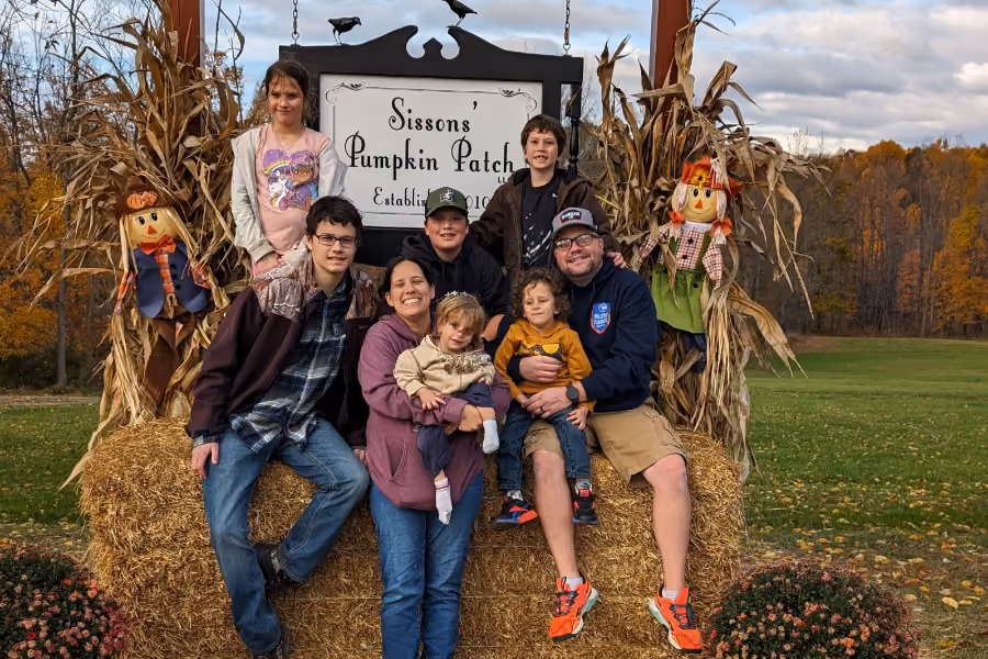 Family of eight posing on hay bales at Sissons' Pumpkin Patch with autumn trees in background.