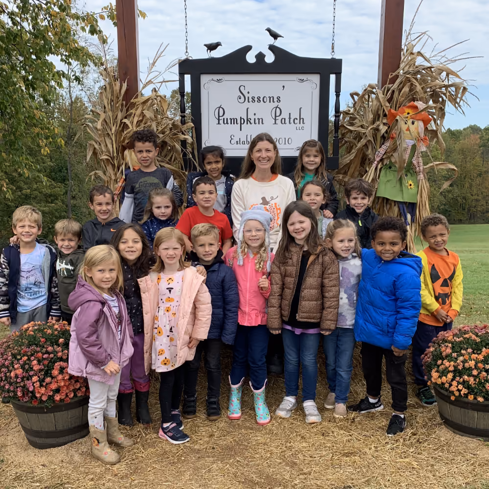 Group of children and one adult smiling in front of a Sissons' Pumpkin Patch sign with fall decorations.