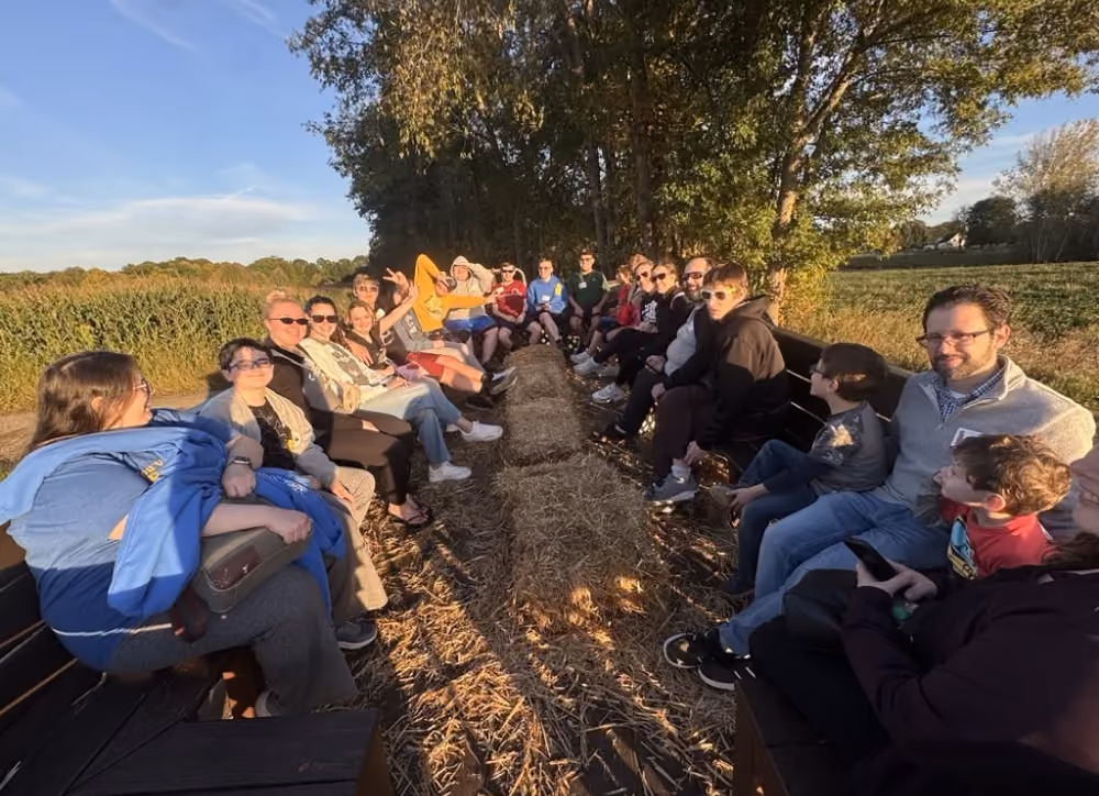 Group of people sitting on benches around hay bales outdoors in a sunny rural area.